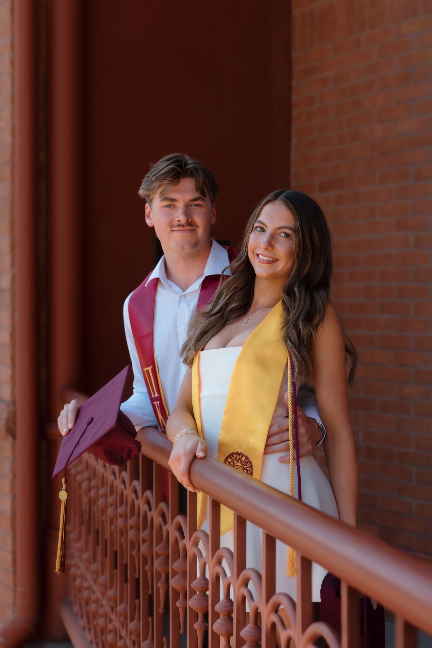 Two smiling graduates holding cap stand together on ornate brick building balcony on sunny day.