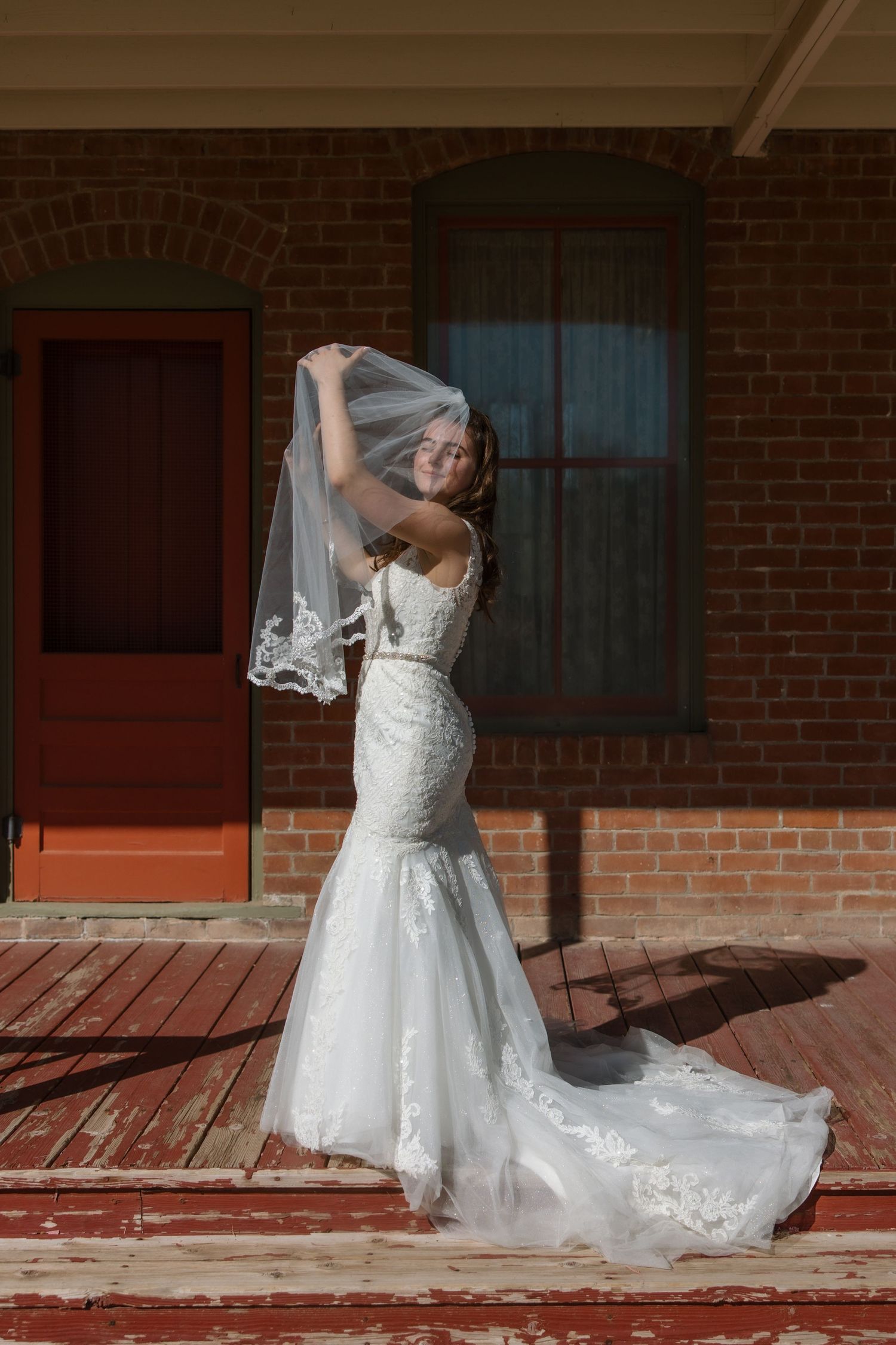 A radiant bride in a fitted lace wedding gown lifts her veil outside a rustic red brick building in sunlight.