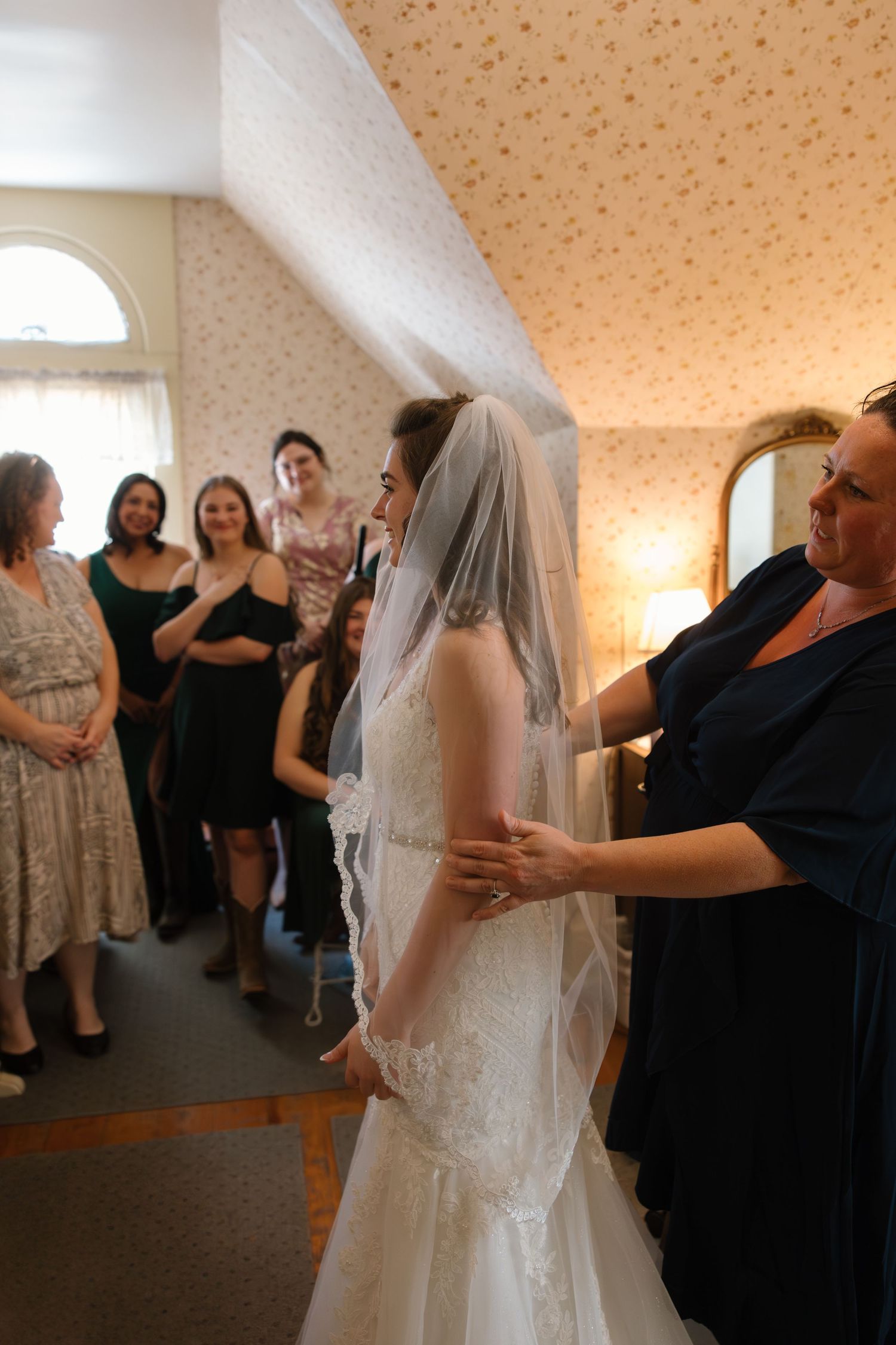 Woman helping bride with veil as guests look on in a floral wallpapered room.