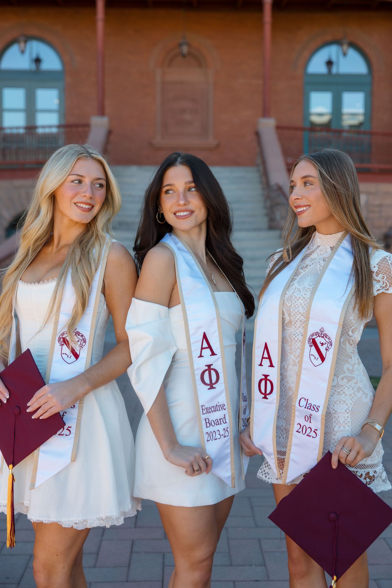 Three smiling women in white dresses with Alpha Phi graduation stoles hold maroon caps outdoors.