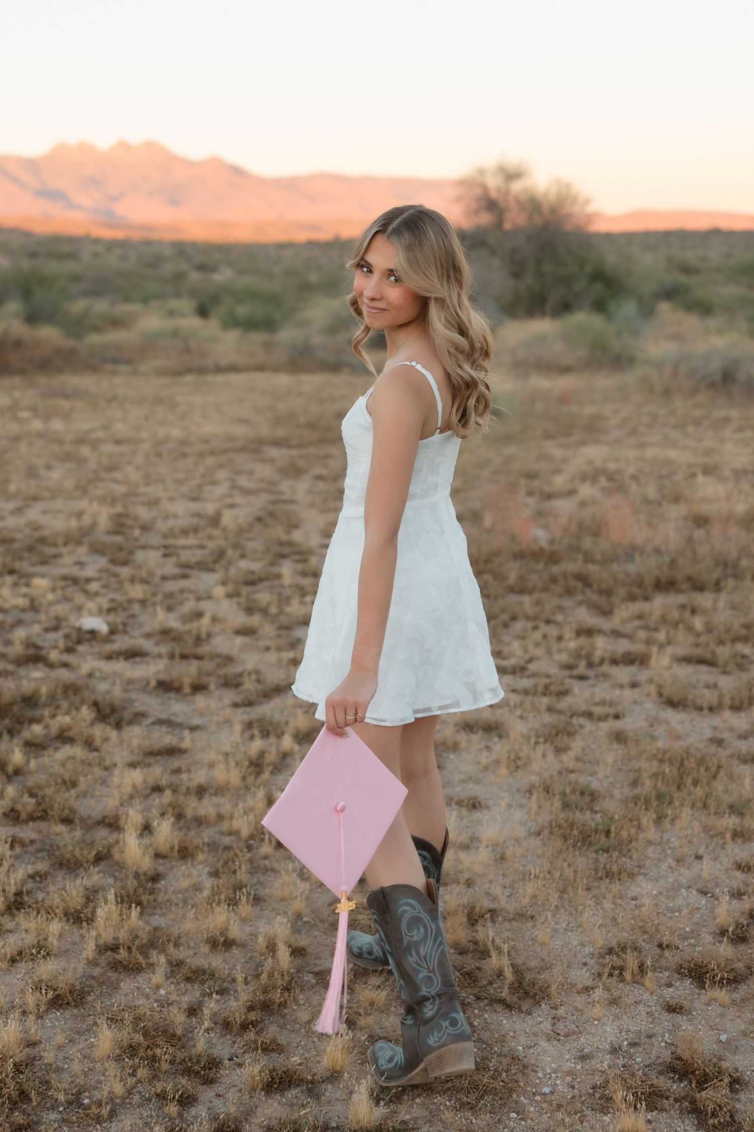 Young woman in white dress and cowboy boots holds pink graduation cap in desert landscape at sunset.