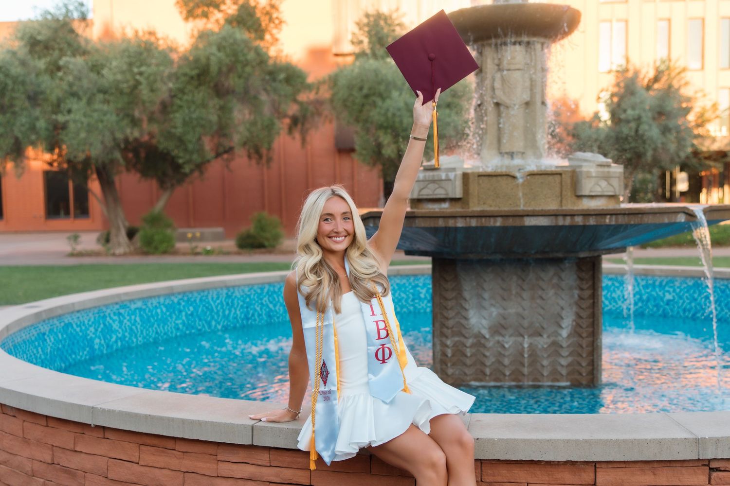 Smiling blonde graduate in white dress tosses cap by fountain, wearing Greek sash in warm sunset light.