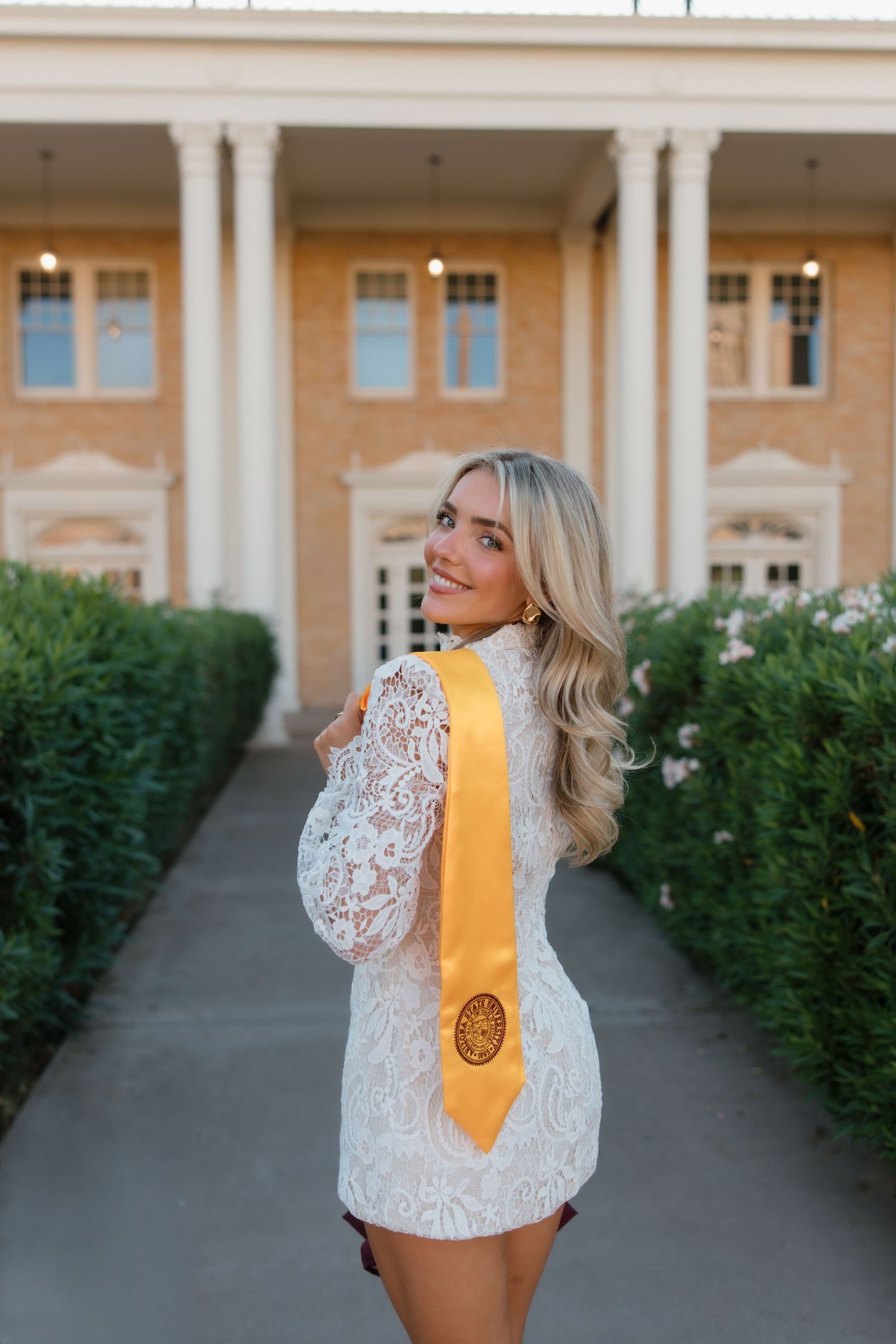 Smiling blonde woman in white lace dress with gold graduation stole poses on pathway outside brick building.