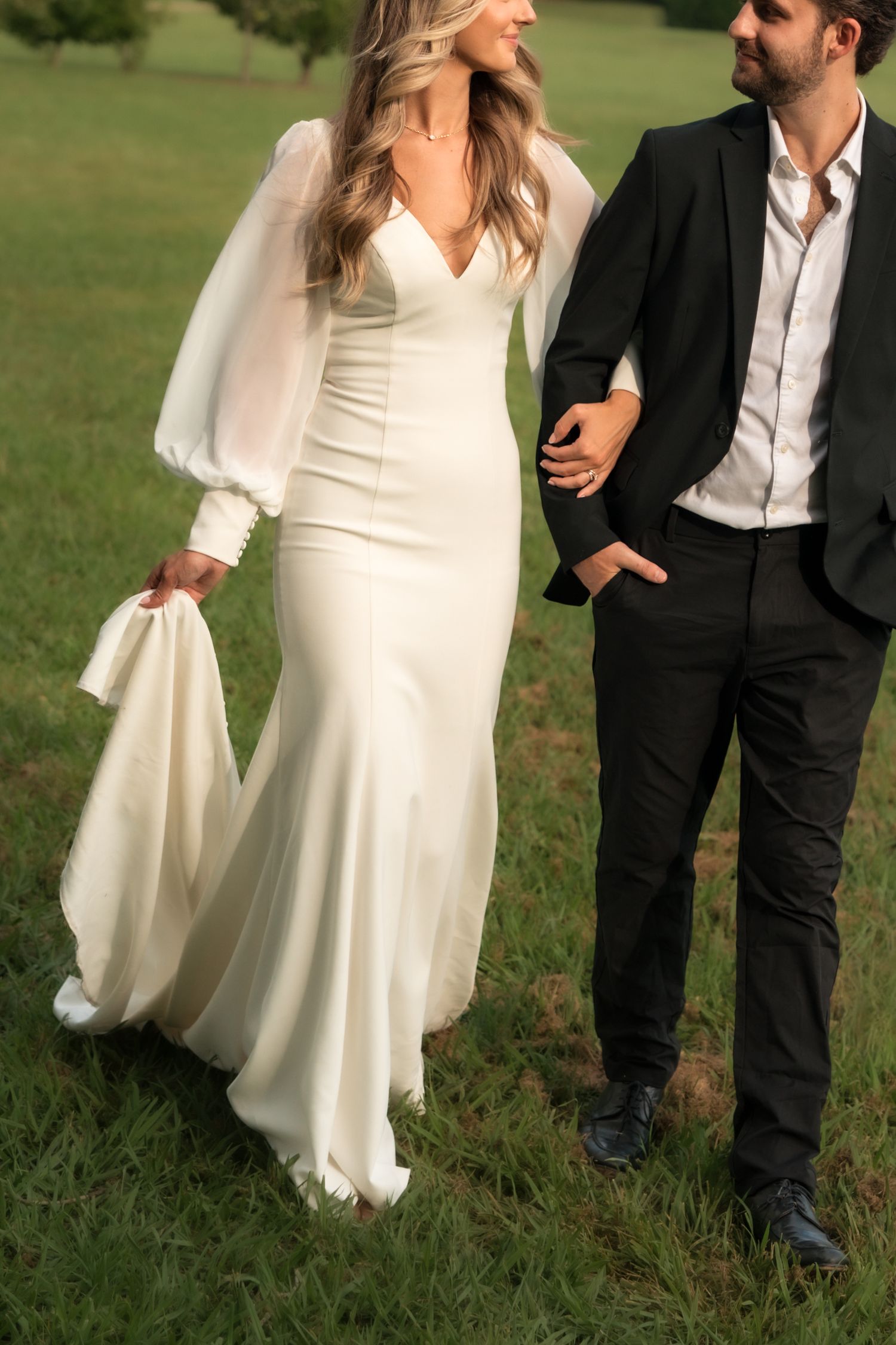 Bride in long-sleeve white mermaid gown holds train while walking hand-in-hand with groom in black suit.