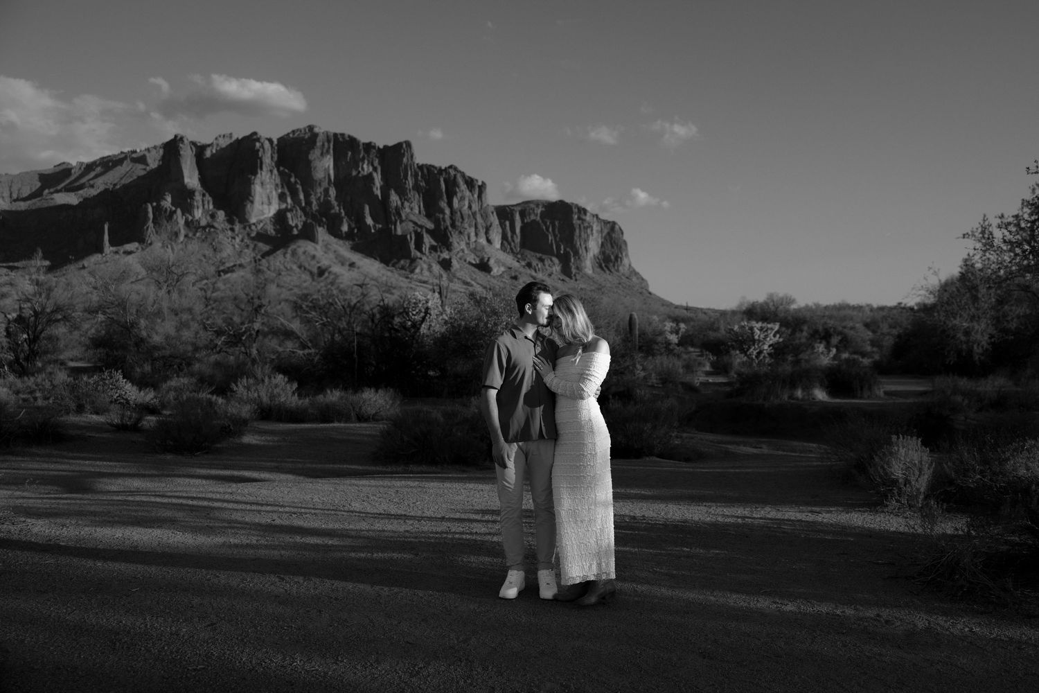 Black and white photo of a couple embracing in a desert landscape with dramatic rocky cliffs in the background.