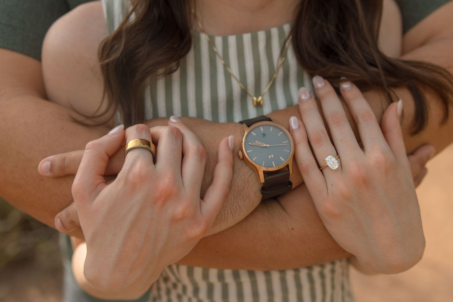 Couple holding hands showing rose gold watch, diamond engagement ring, gold wedding band, and layered necklace.