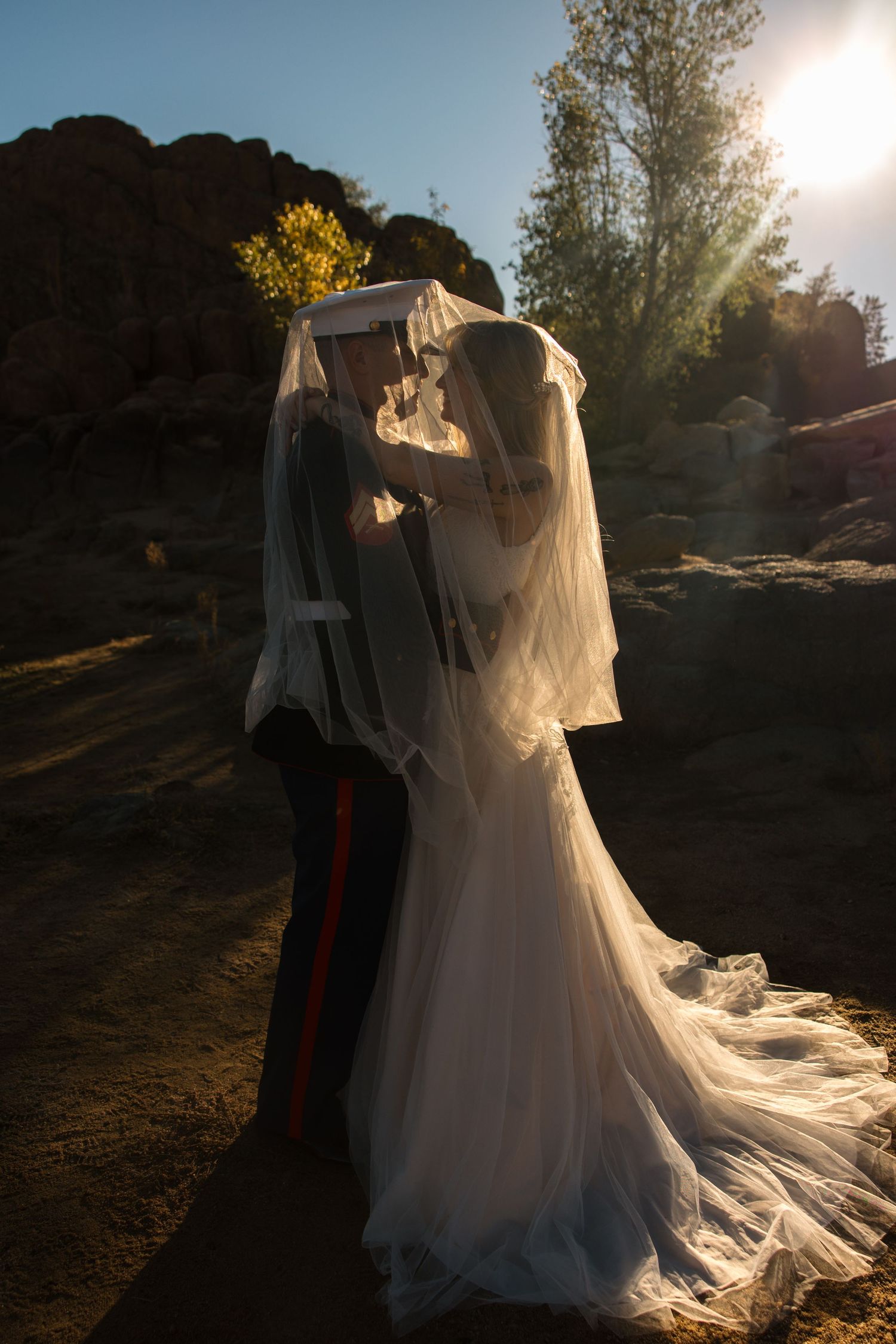 Marine groom and bride kiss under veil with sun flare in desert landscape.