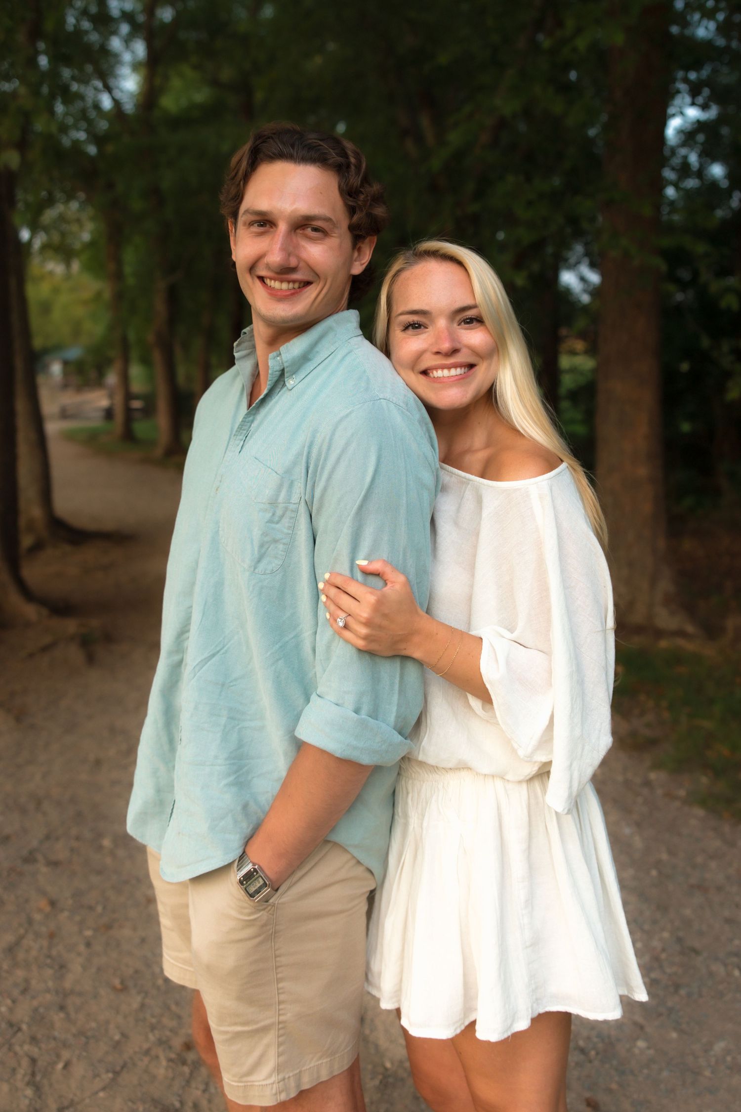 Happy couple hugging and smiling on a tree-lined path, wearing casual summer outfits in natural light.