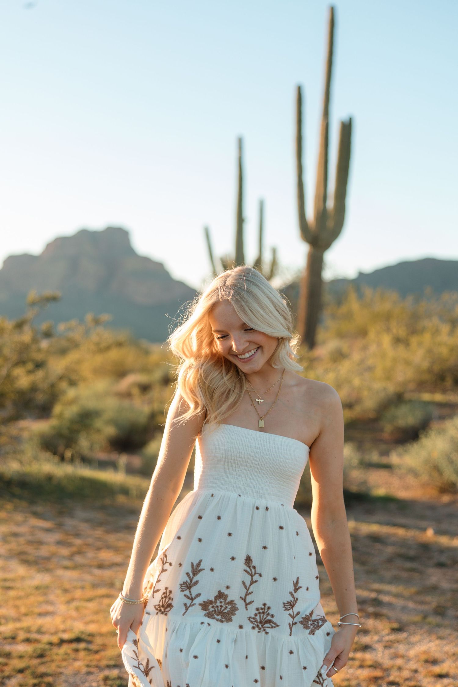 Blonde woman in white floral strapless maxi dress laughing in Arizona desert at golden hour.