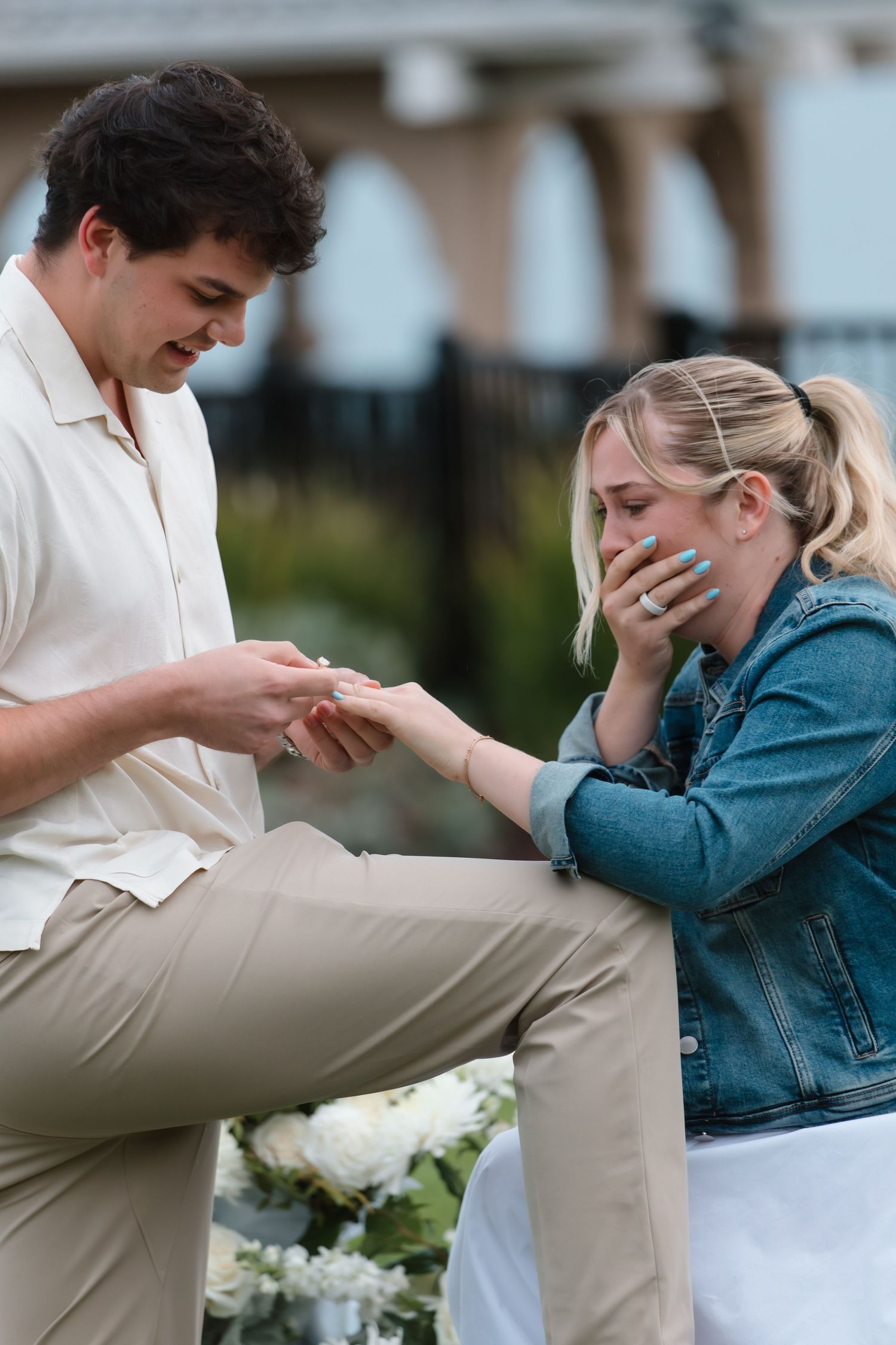 Emotional woman covers mouth as man kneels placing engagement ring on her finger amid white floral arrangements.