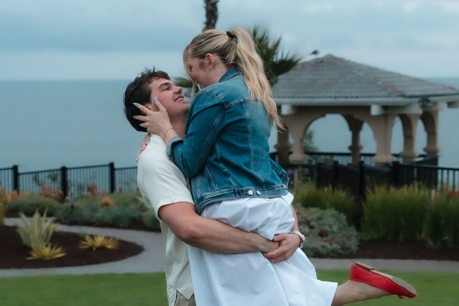 A couple shares a romantic embrace outdoors, with the man lifting the woman near a scenic waterfront gazebo.