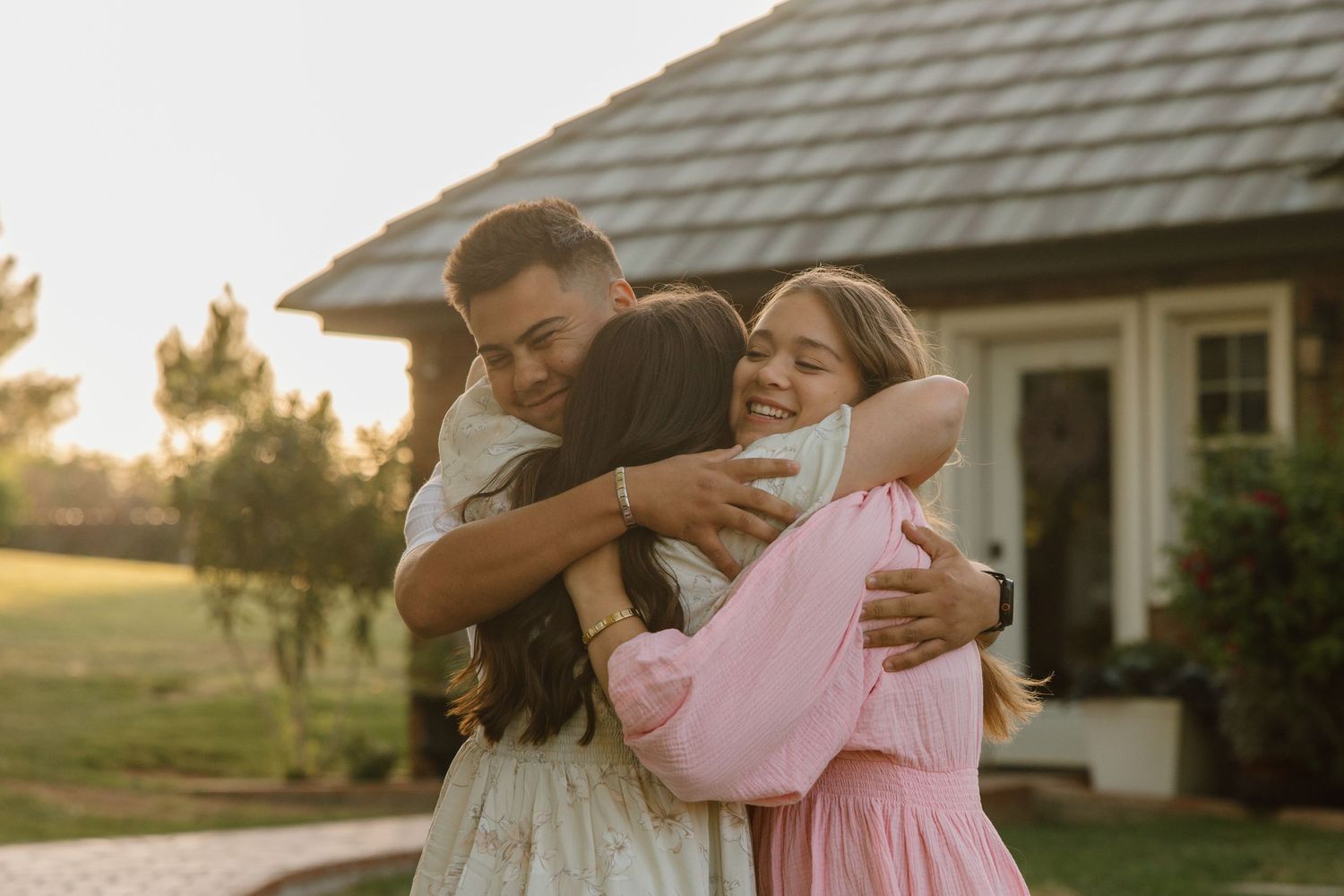 A joyful group of three people sharing a warm embrace outdoors near a house during golden hour sunset.