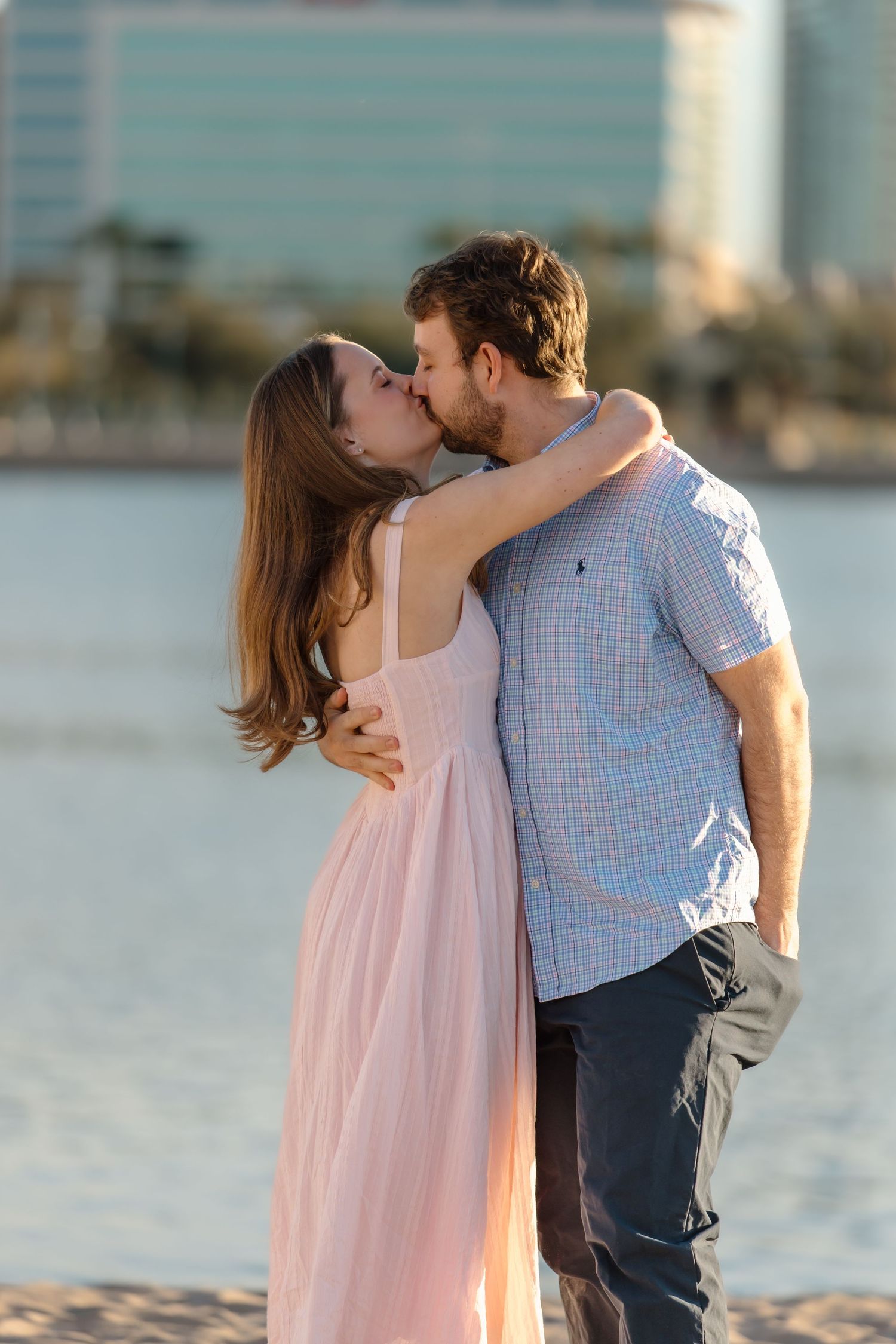 Couple sharing a romantic kiss on a sandy beach at sunset, woman in pink dress, man in blue plaid shirt.