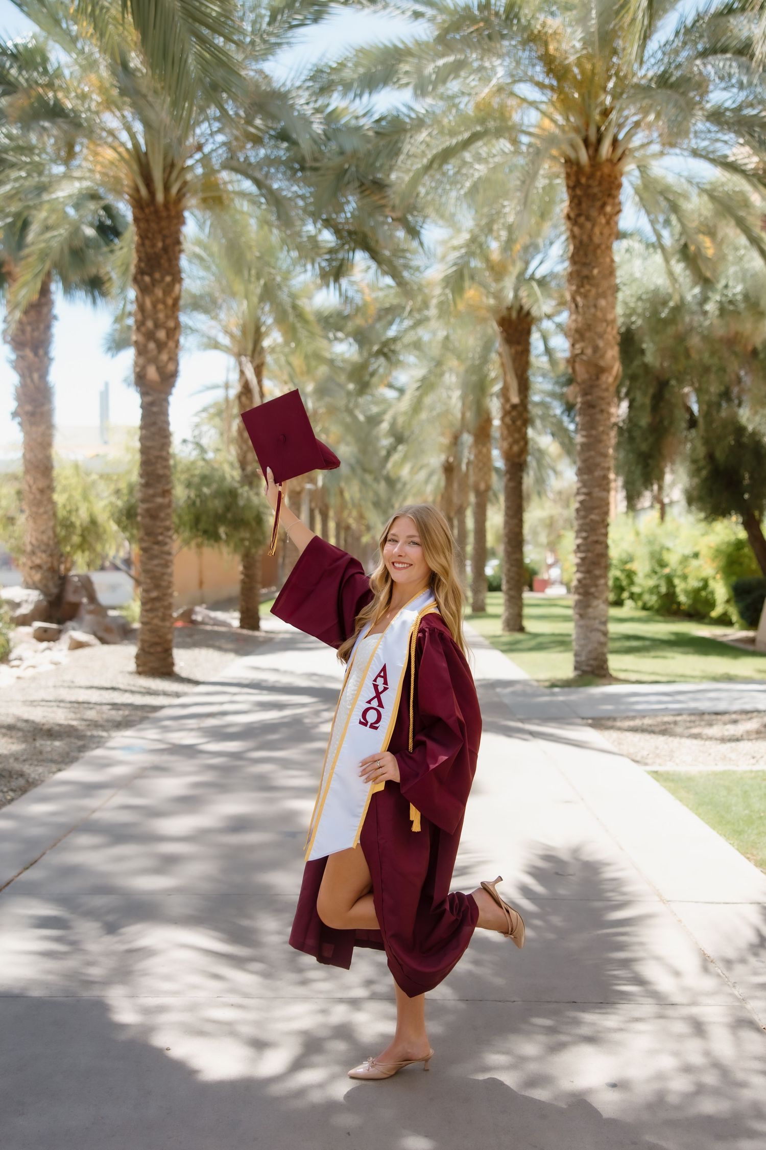 A smiling graduate in maroon cap and gown celebrates outdoors among tall palm trees, joyfully raising her cap.