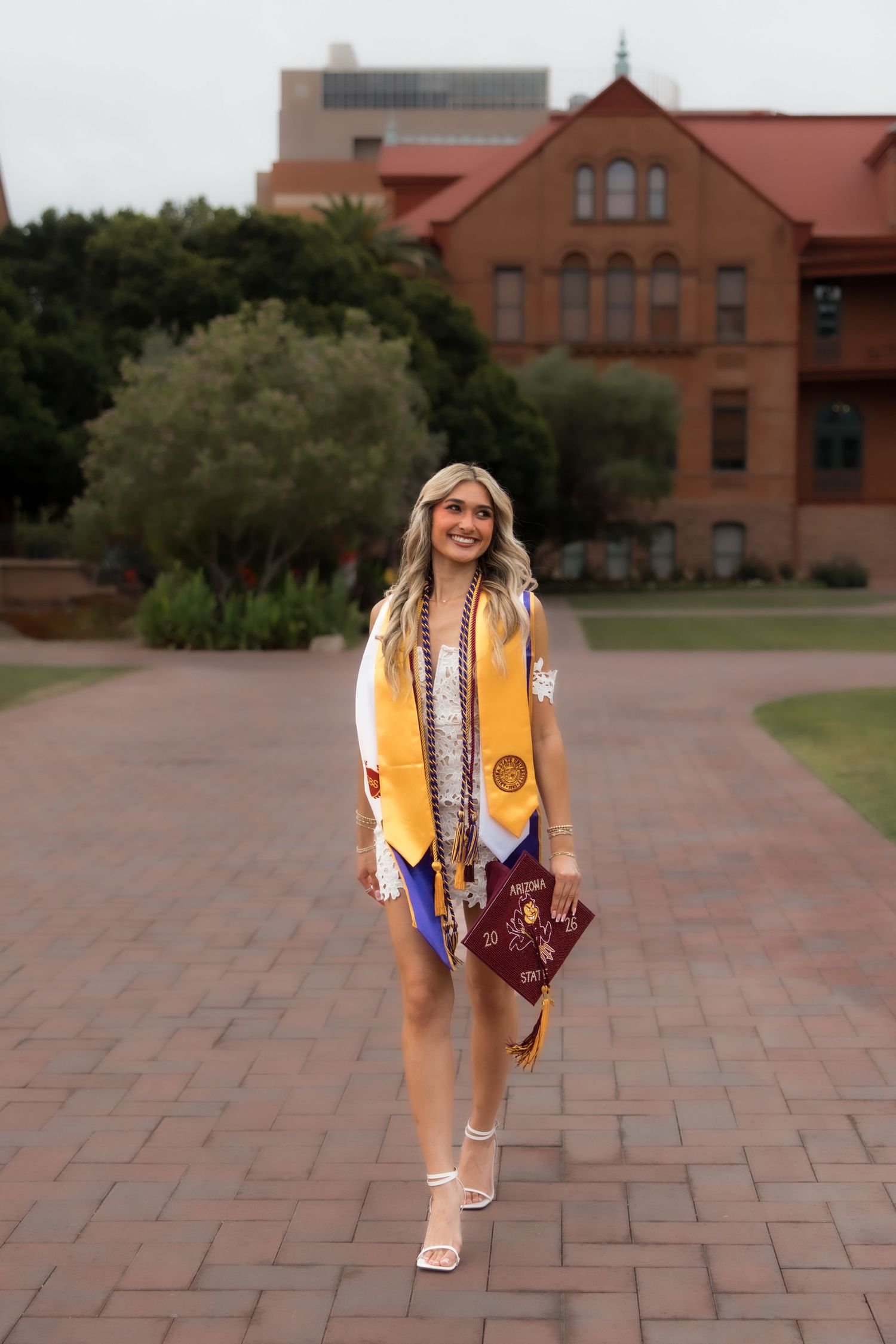 Smiling female graduate wearing gold stole holds diploma on brick campus path with red brick building background.