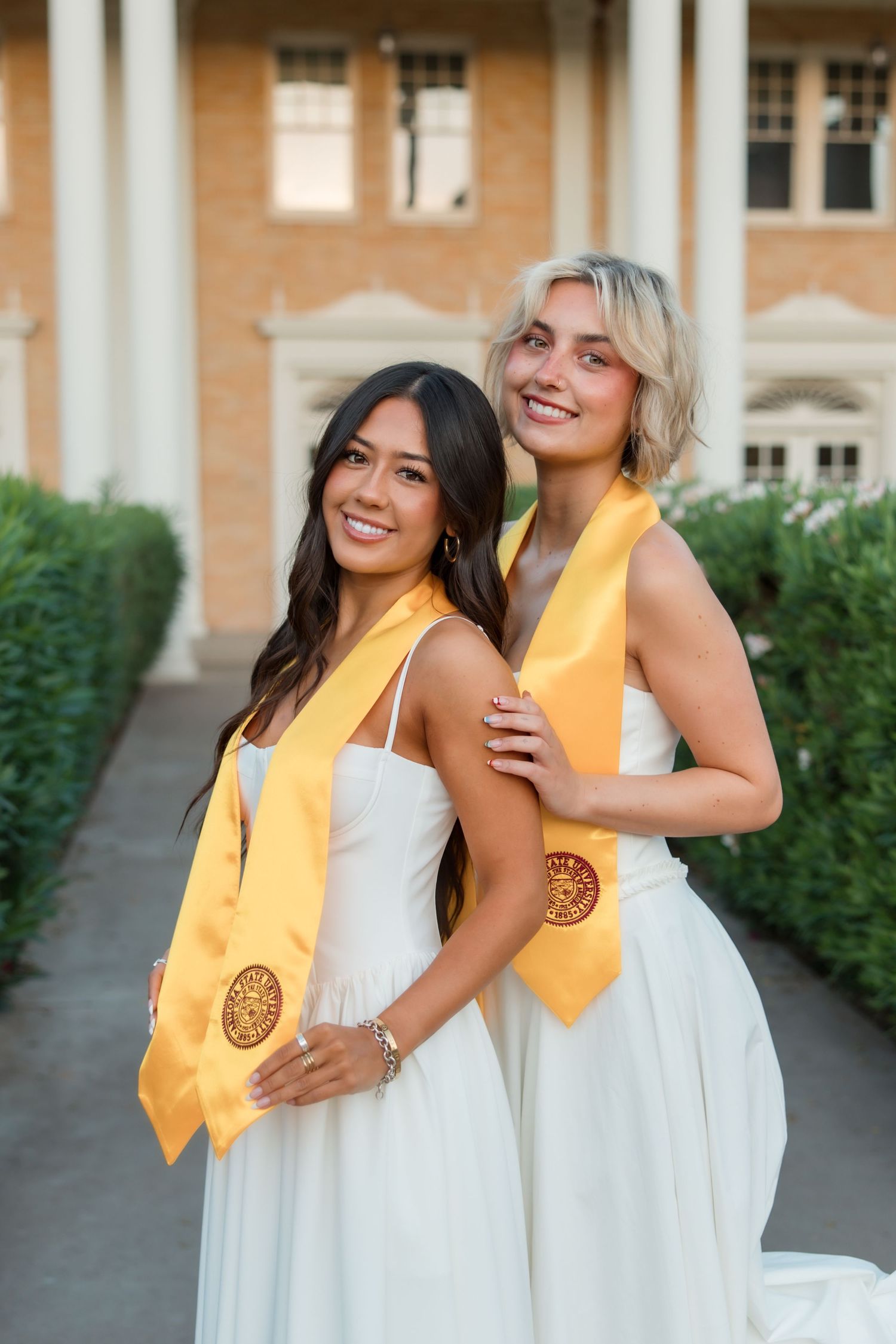 Two smiling female graduates in white dresses wearing gold graduation stoles pose together outdoors near a brick building.