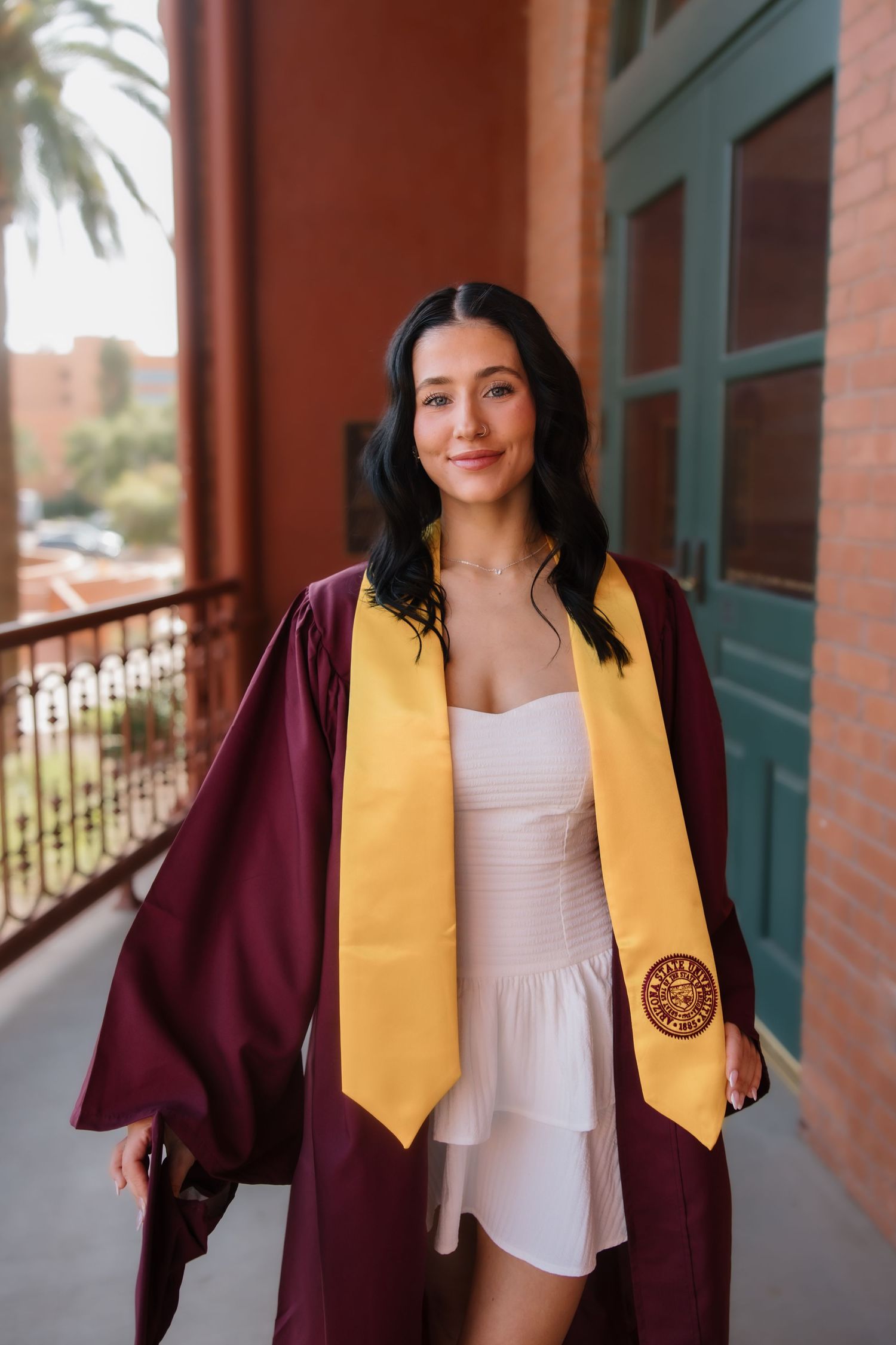 Young woman in maroon graduation gown with gold stole smiles on a balcony, celebrating her academic achievement.