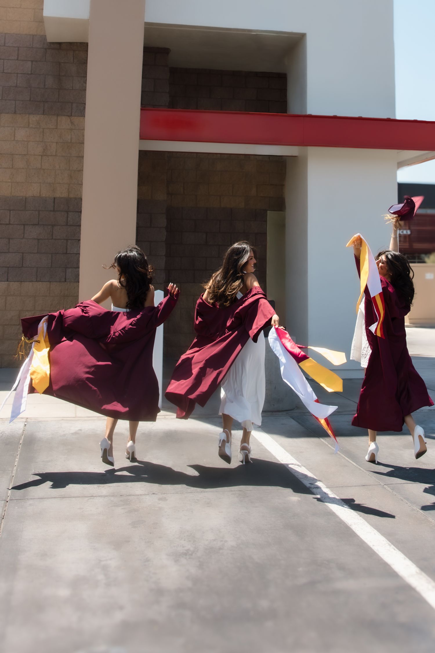Three graduates in maroon caps and gowns running joyfully outside a building.