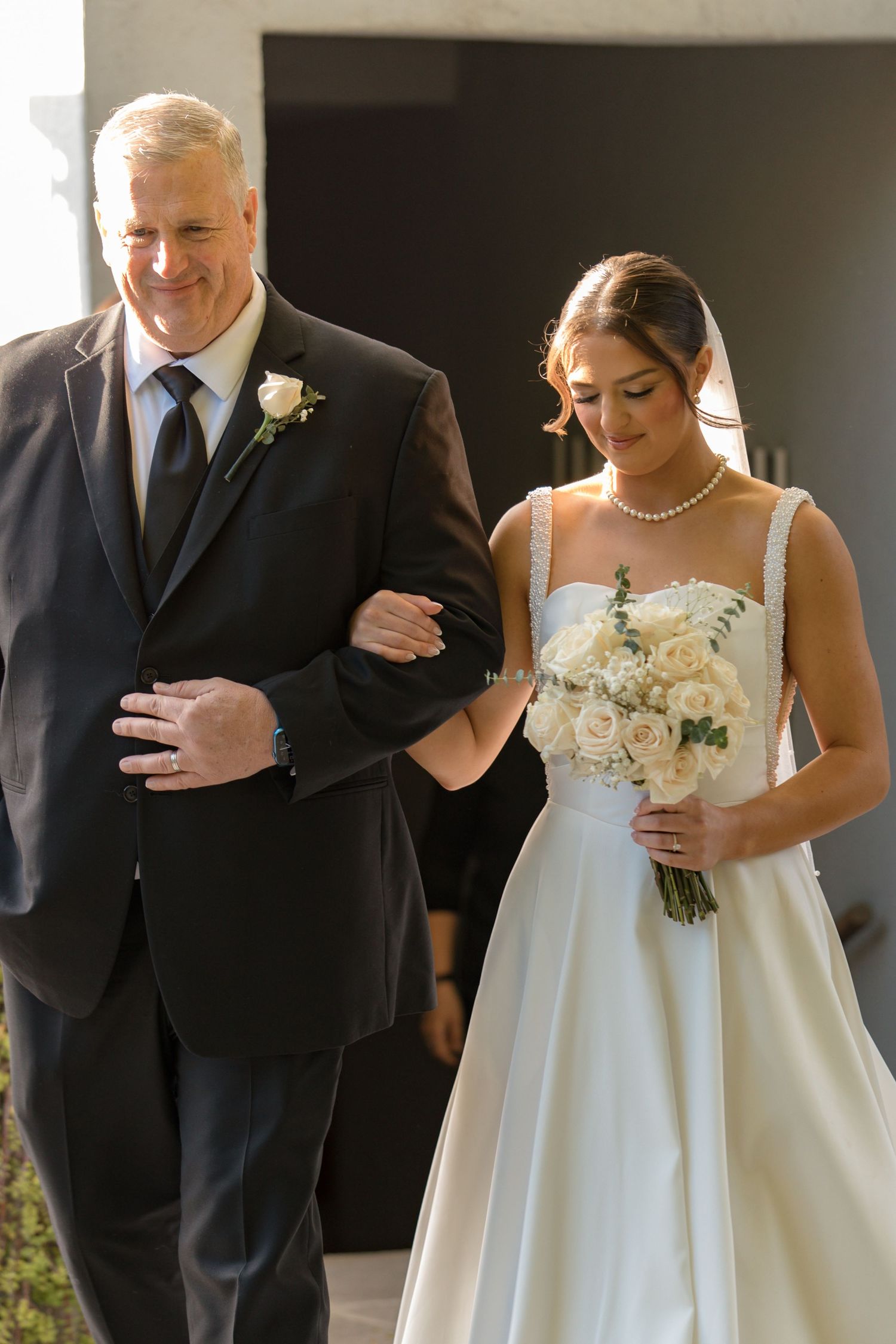 Bride in white gown holding rose bouquet walks down aisle with father in black suit.