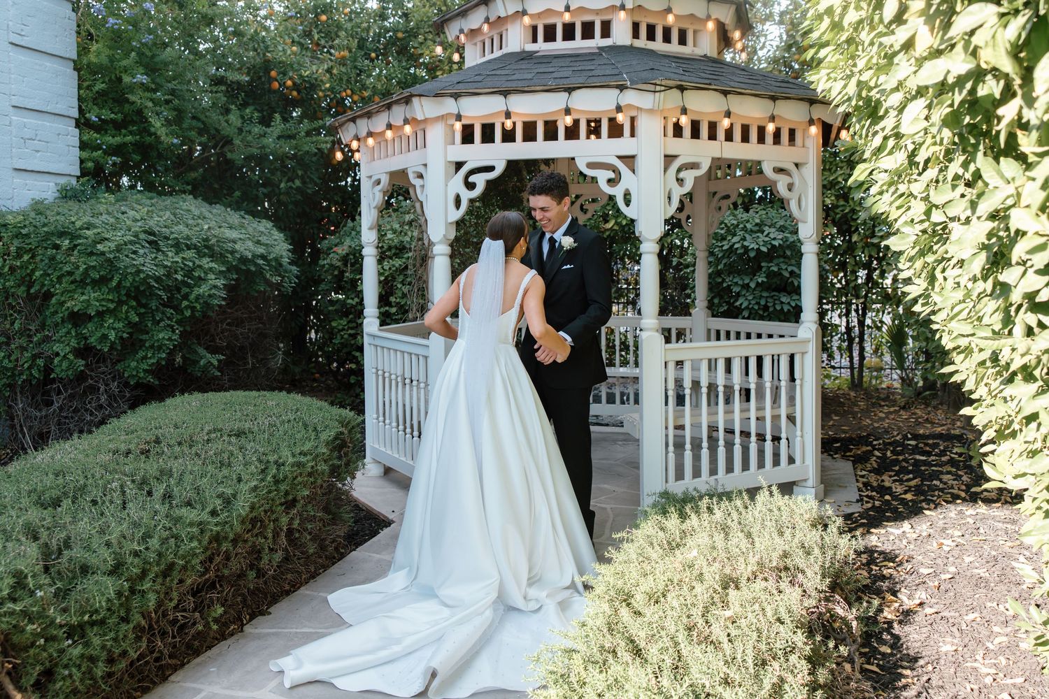 Bride and groom hold hands and kiss beside a white garden gazebo surrounded by lush greenery.