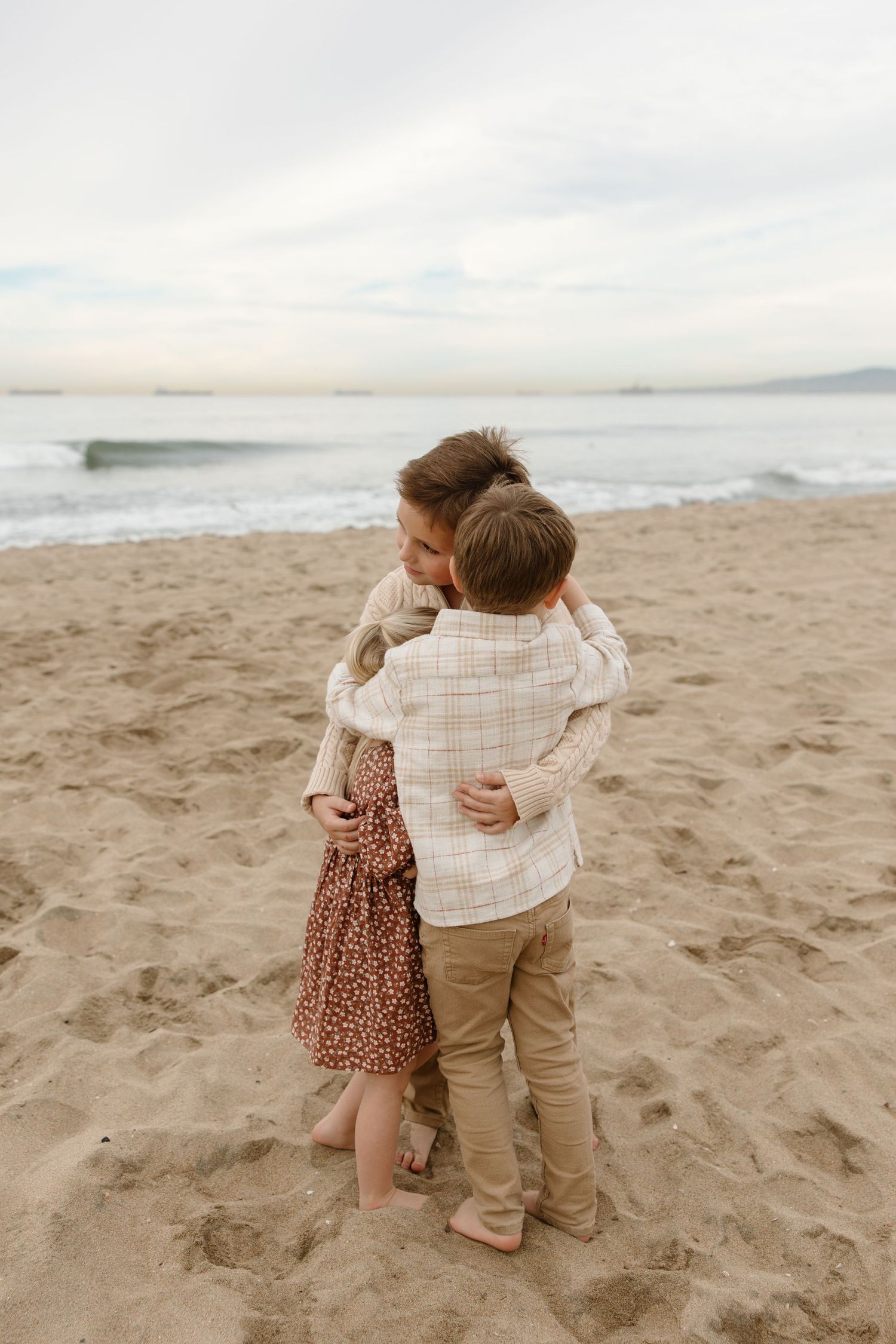 Two young children share a tender hug on a sandy beach with ocean waves in the background.