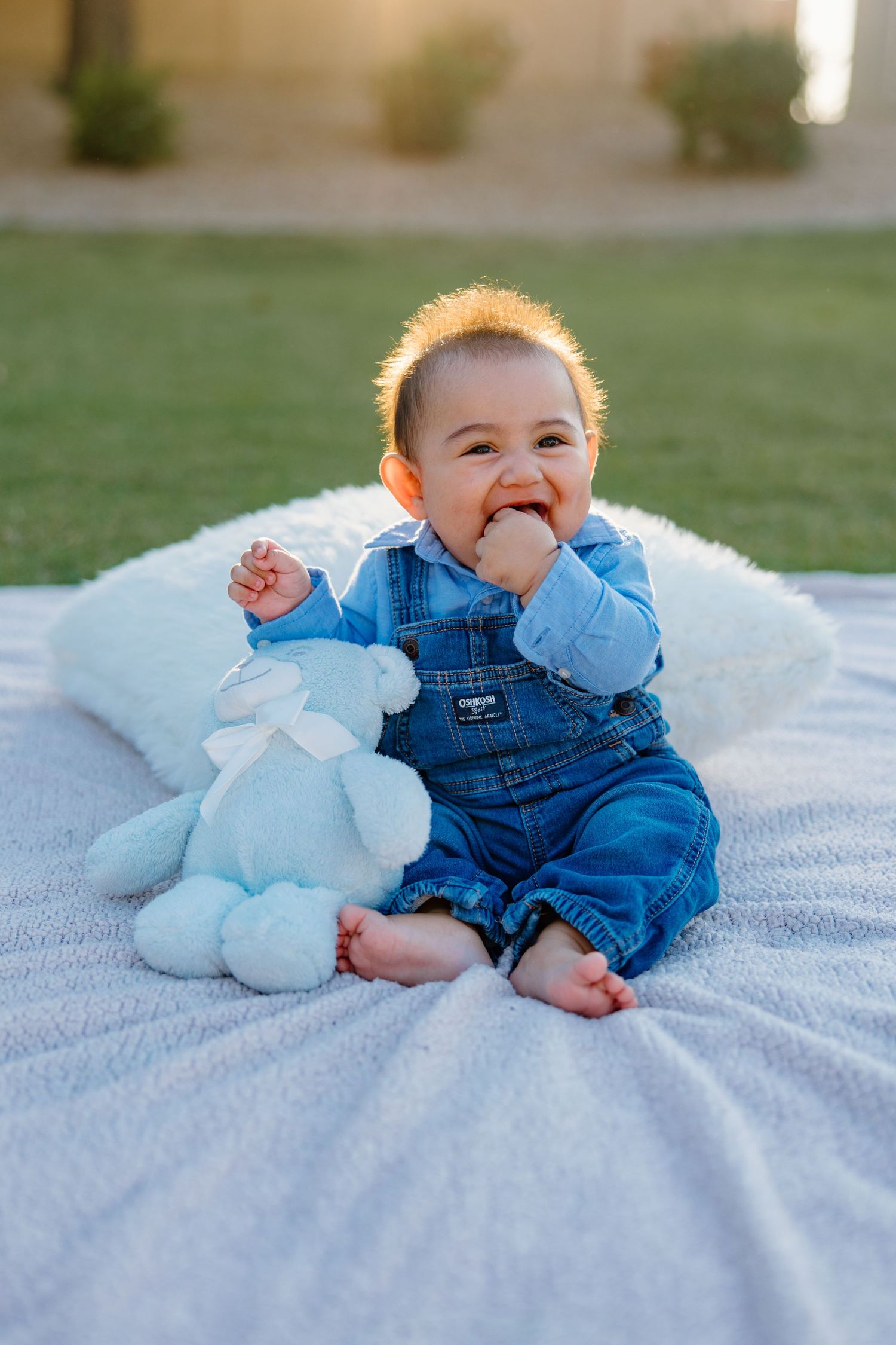 Smiling baby in denim overalls sitting on a white blanket outdoors with a blue stuffed animal toy.
