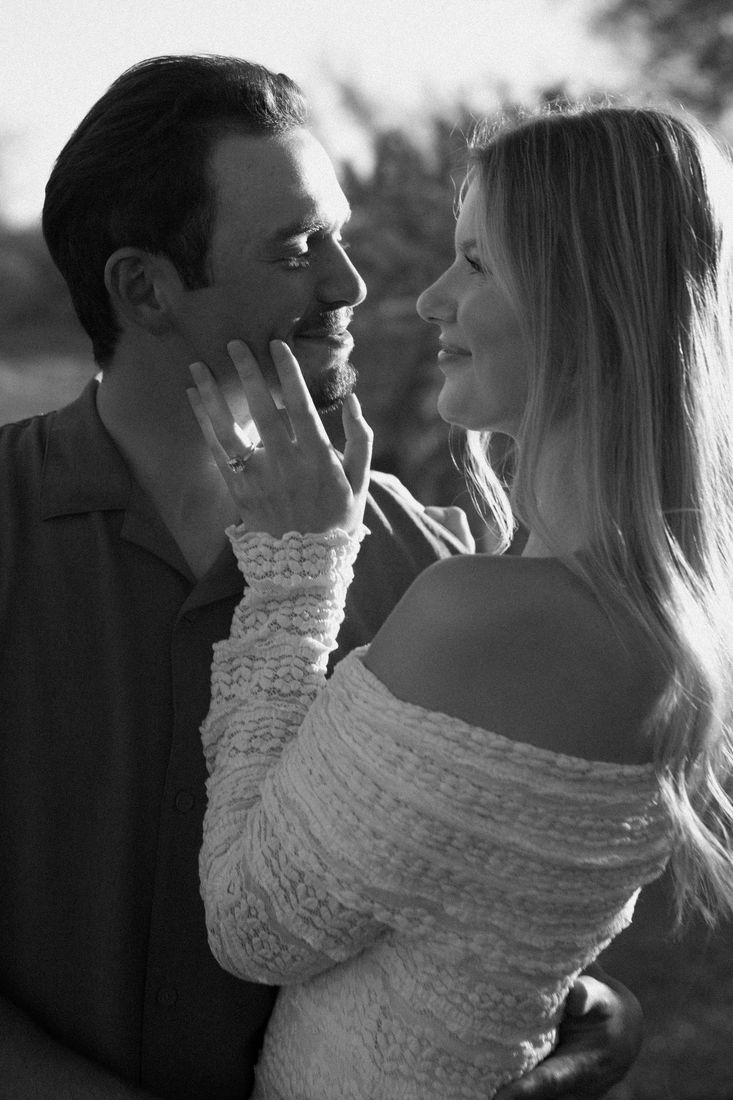 Black and white photo of a smiling couple, woman showing off engagement ring while gazing at her partner.