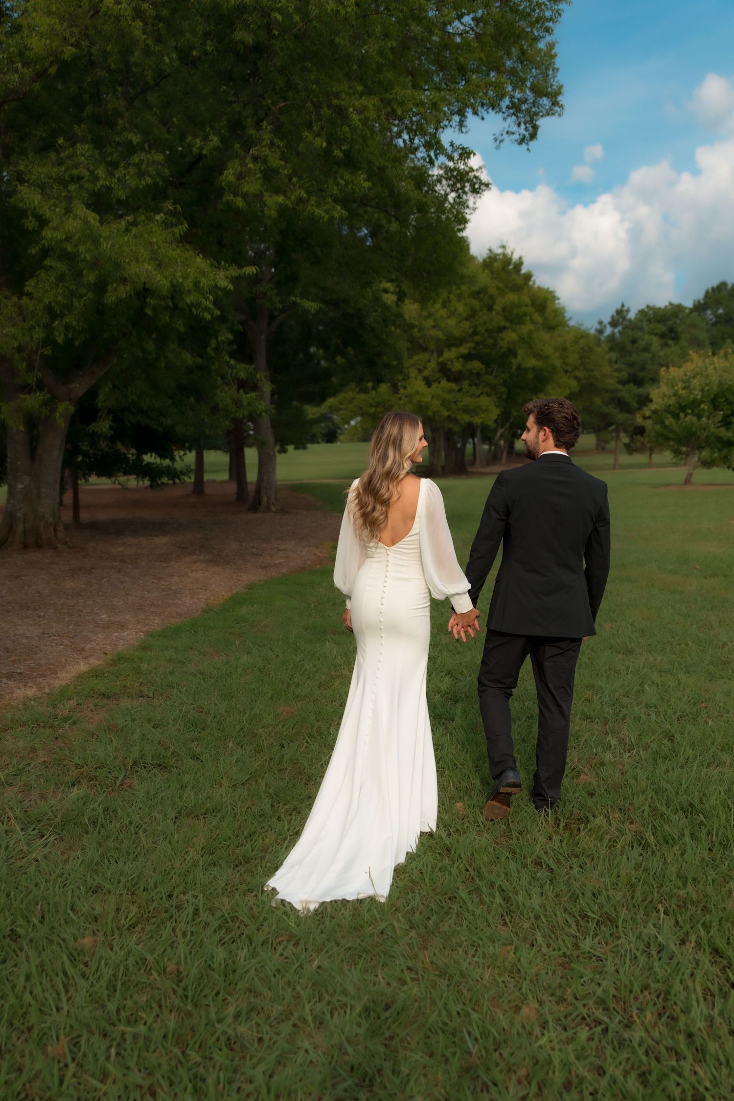 Bride and groom walk hand in hand through lush green park, bride wearing elegant white backless gown.