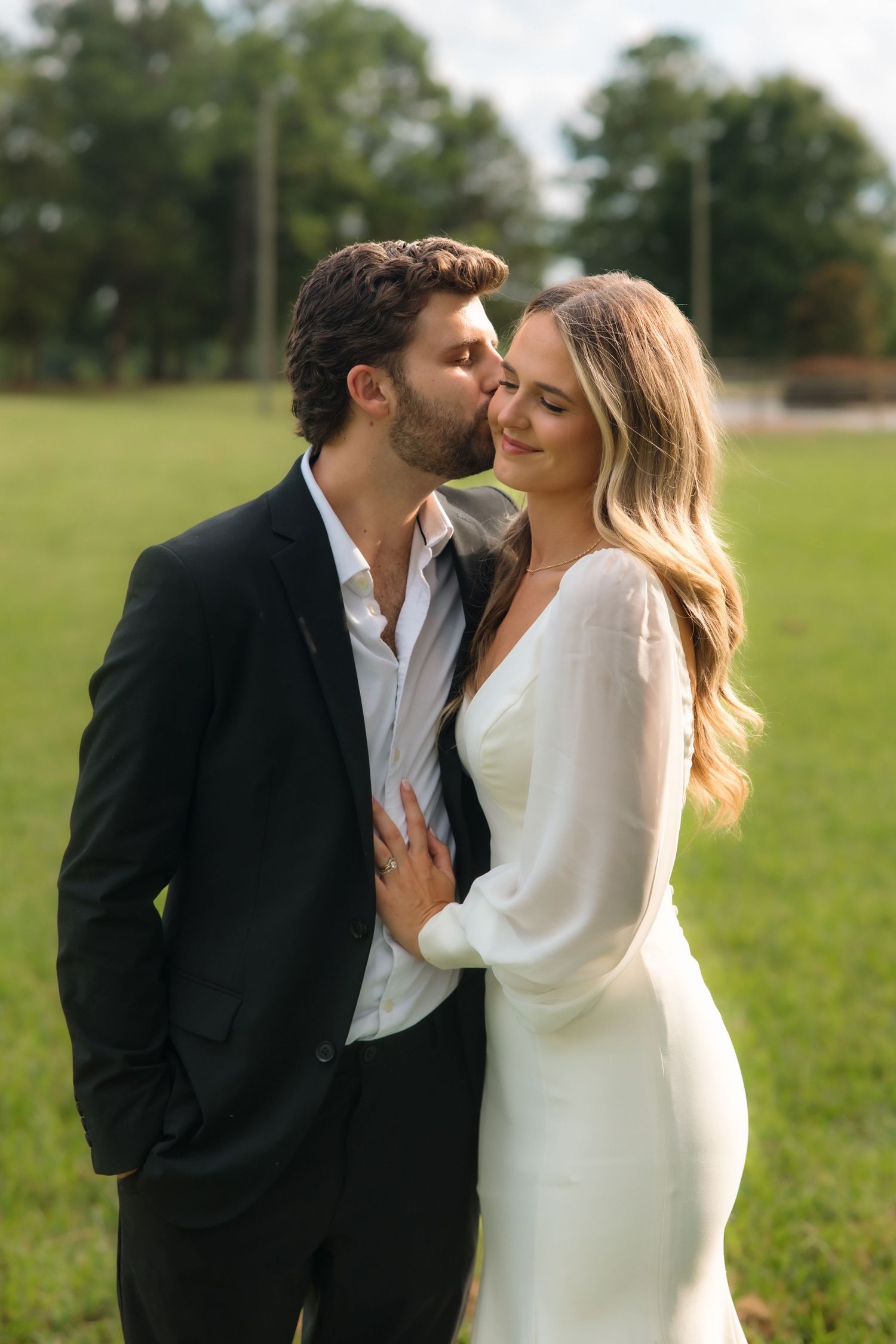 Groom in black suit kissing bride on forehead in white dress outdoors on green grass.