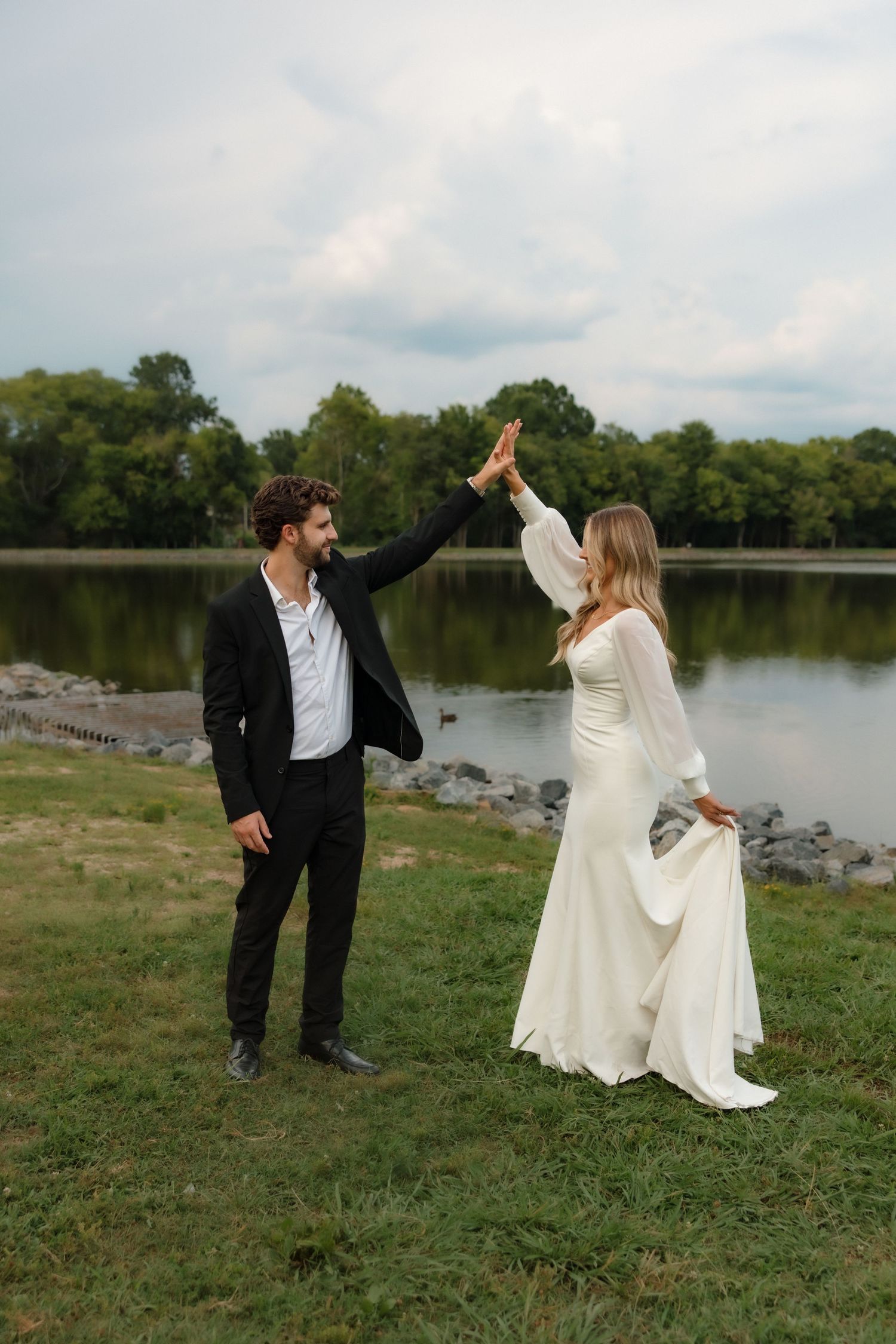 Joyful bride and groom high-fiving outdoors by a lake, dressed in white gown and black suit.