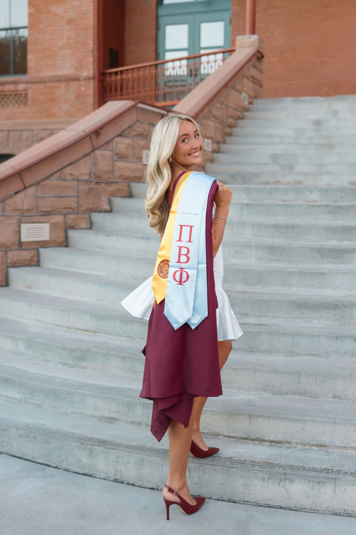 Blonde woman in maroon dress wearing Pi Beta Phi graduation sash posing on brick steps.