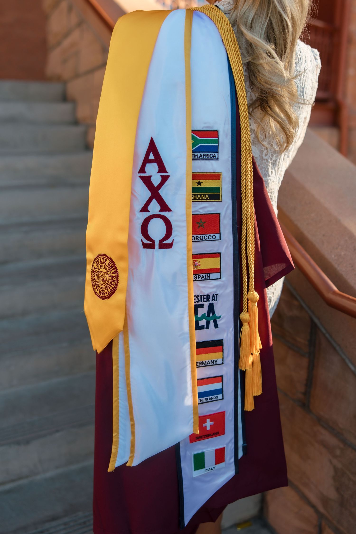 Graduation stole with Alpha Chi Omega letters and international country flags displayed on white and gold sash.
