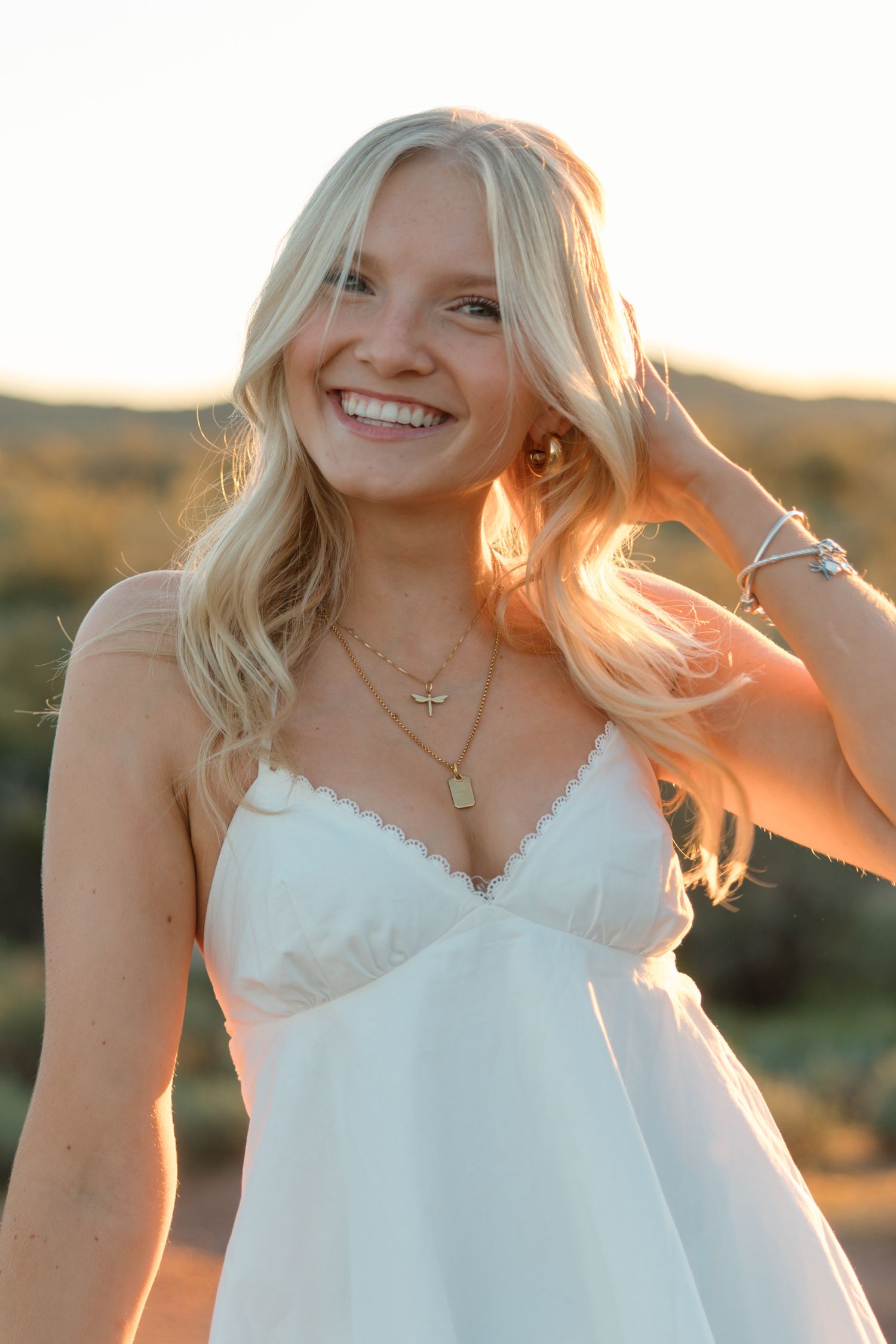 Smiling blonde woman in white dress with gold necklaces, posed outdoors in golden hour sunlight.