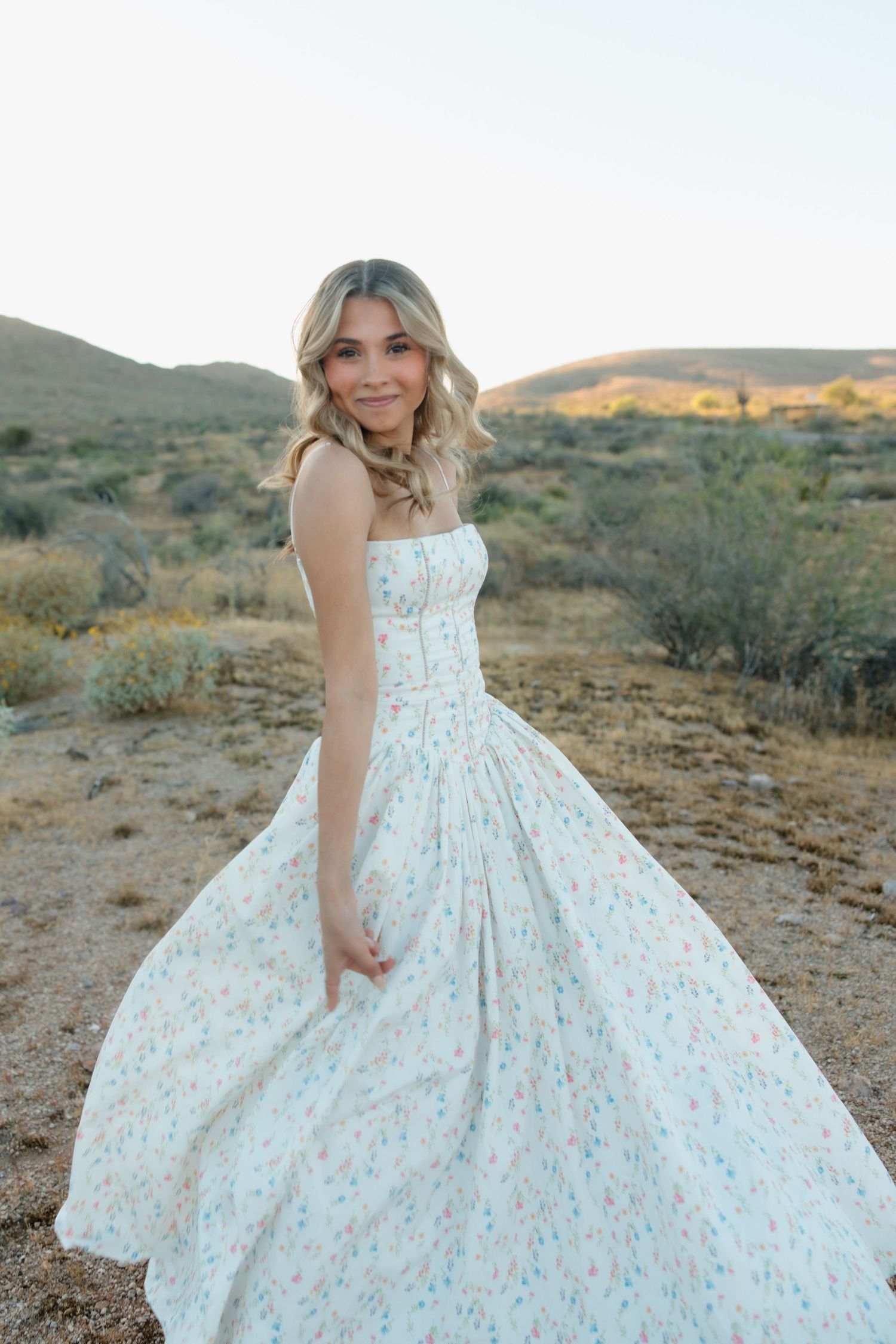 A woman in a floral white maxi dress poses gracefully in a desert landscape at dusk.