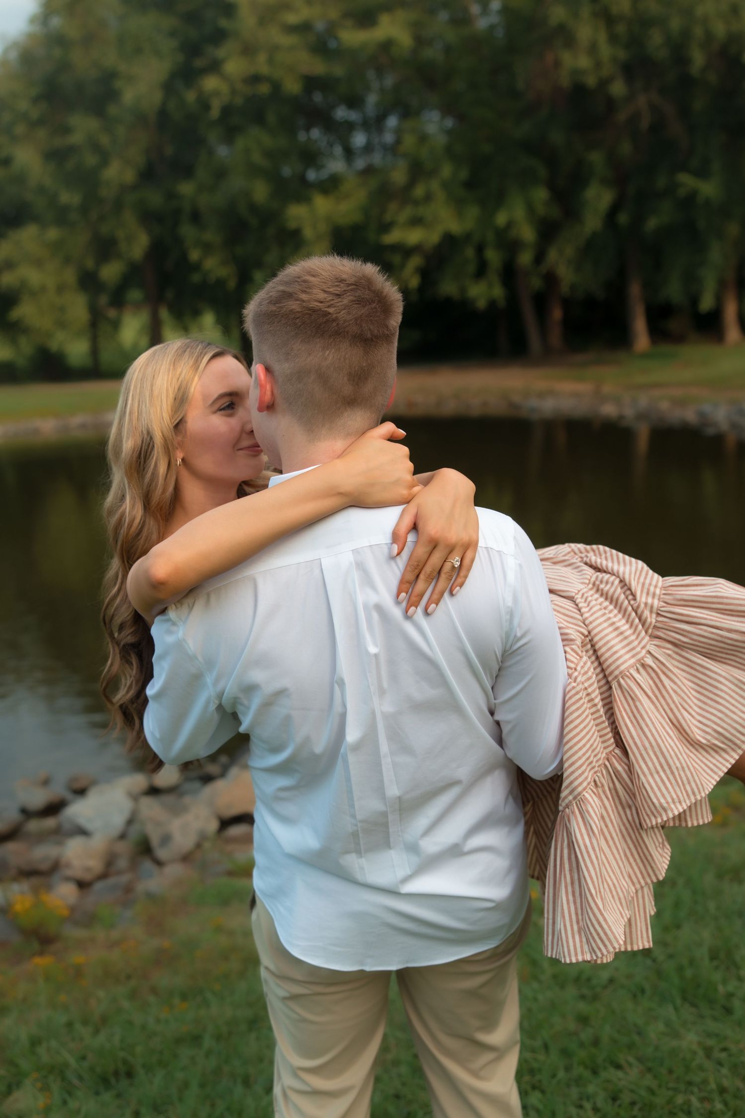 A couple embraces near a pond in a park, the woman in a striped skirt hugging a man in a white shirt.