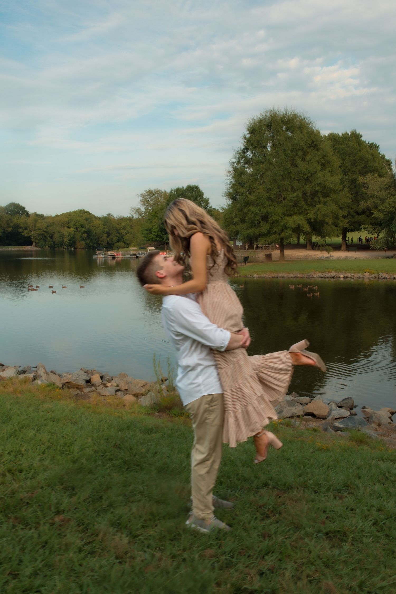 A man lifts a woman for a romantic kiss by a serene lake in a lush green park.