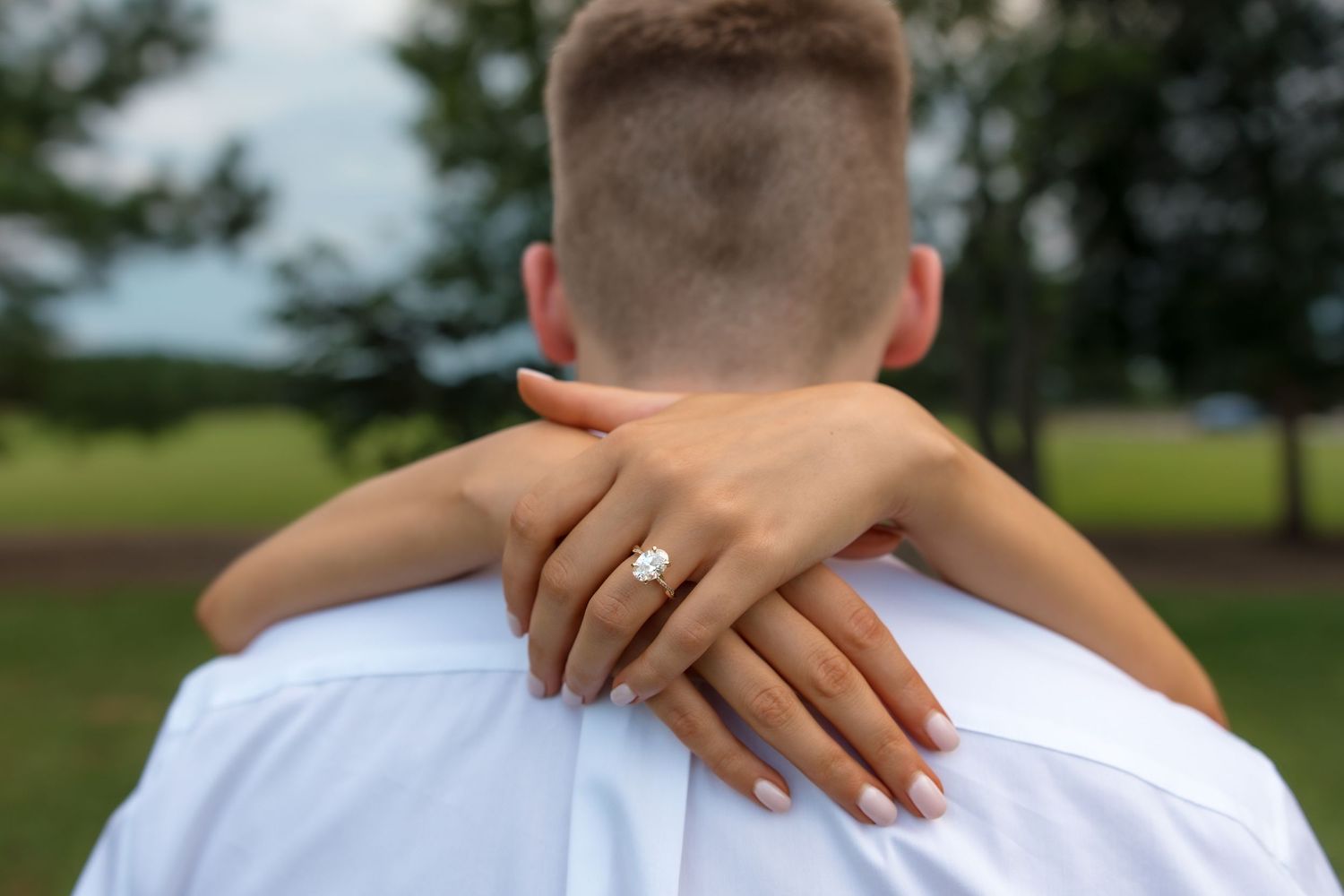 Woman wearing diamond engagement ring embracing man from behind outdoors in a romantic proposal moment.