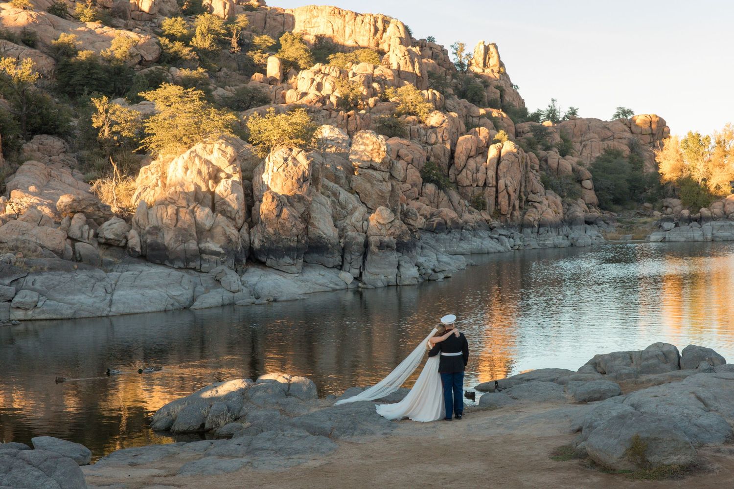Bride and Marine groom share romantic moment by reflective lake with dramatic rocky cliffs.