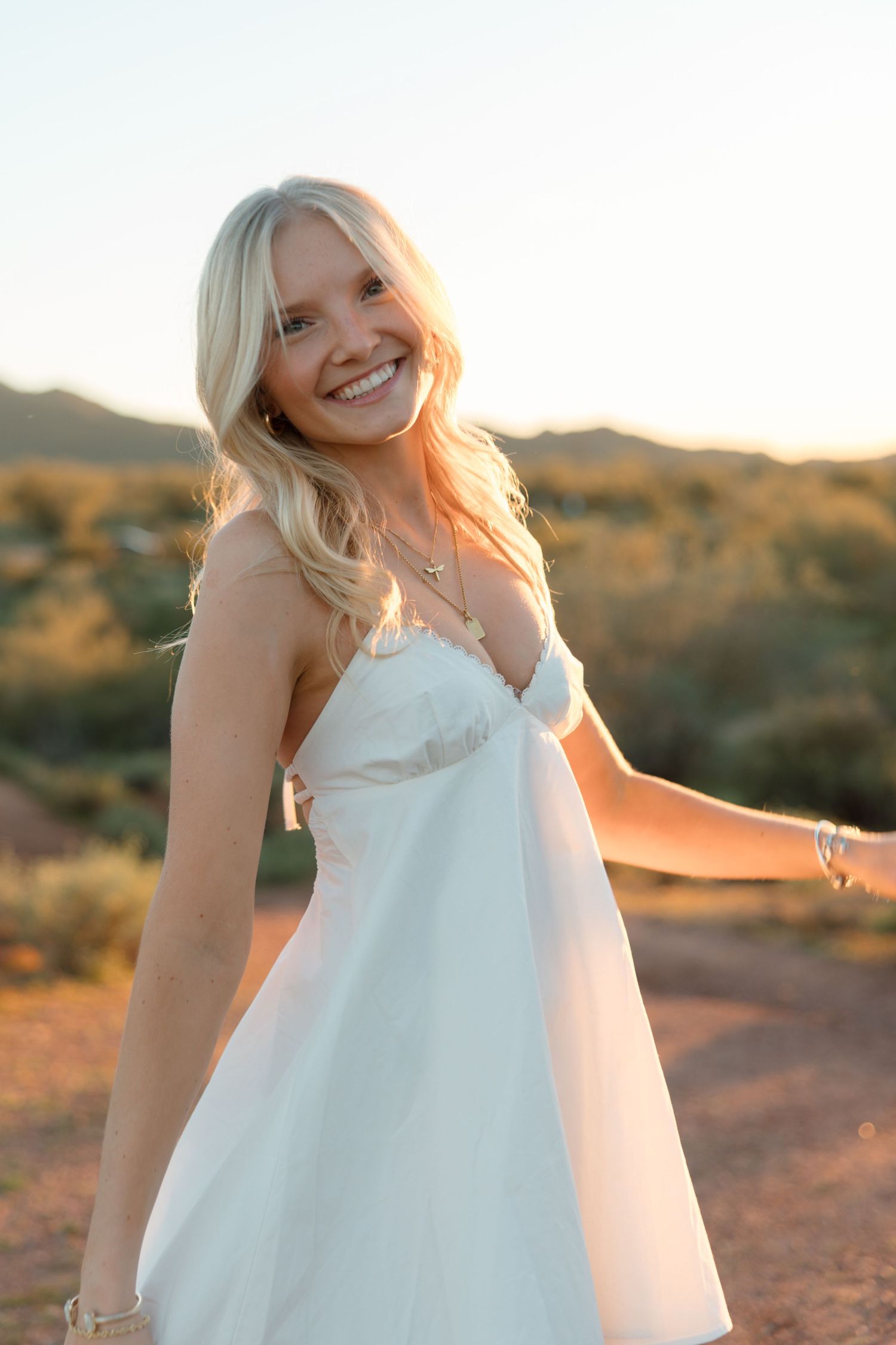 Cheerful blonde woman in white sundress spinning in warm golden sunset desert scene.