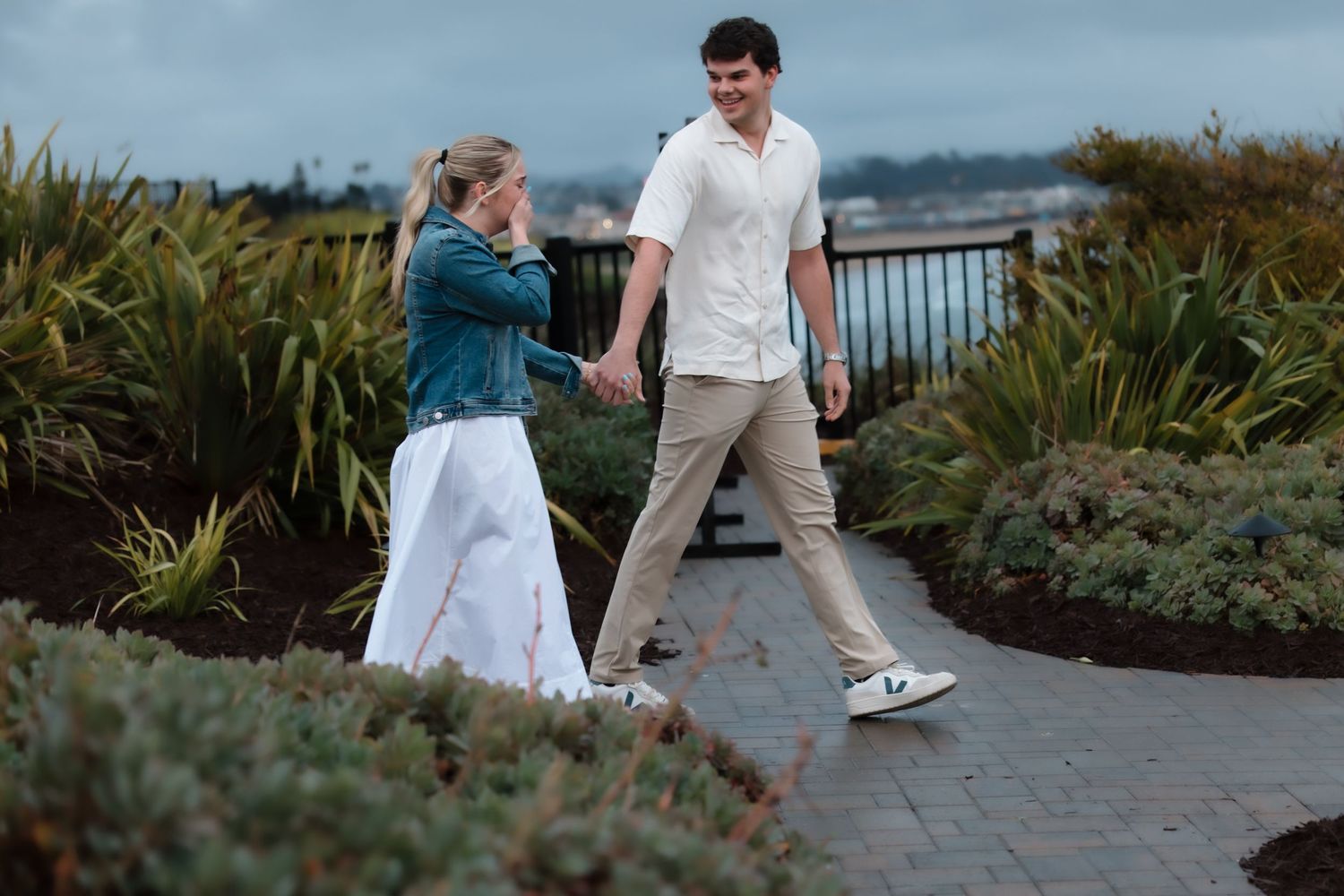 Couple holding hands walking on a brick path surrounded by lush greenery near a waterfront on a cloudy day.