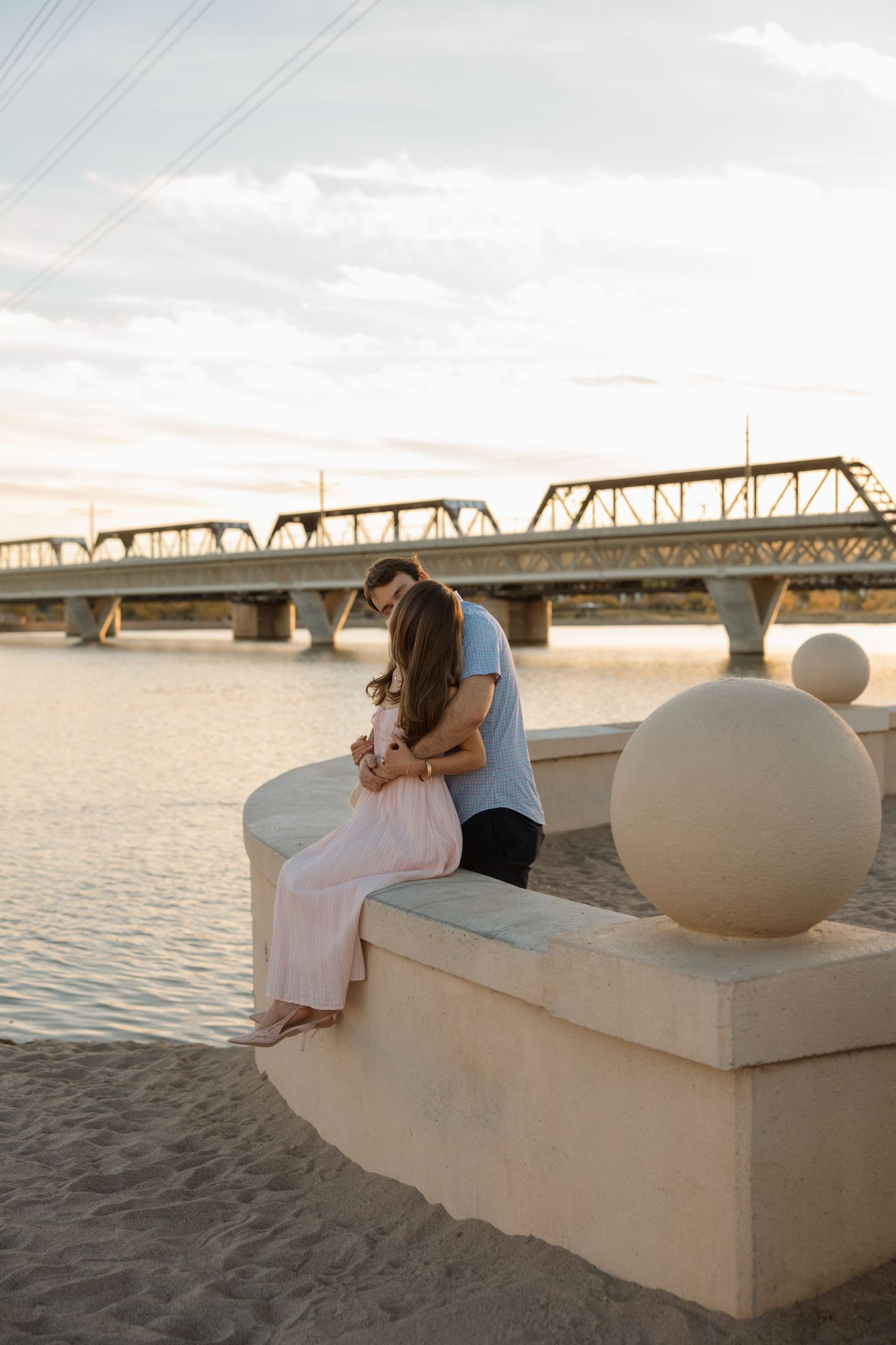 Romantic couple sitting close together on stone wall by river at golden hour with bridge in background.