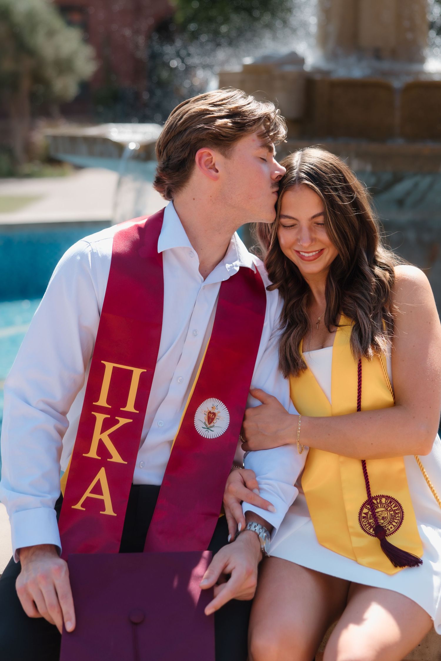 Couple in graduation stoles, man wearing Pi Kappa Alpha sash kisses woman's head near fountain.