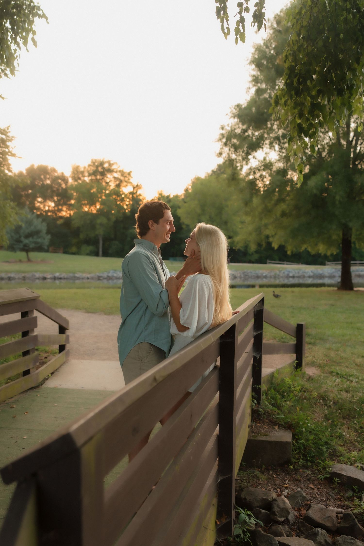A couple shares a romantic moment on a wooden bridge at sunset in a lush green park.