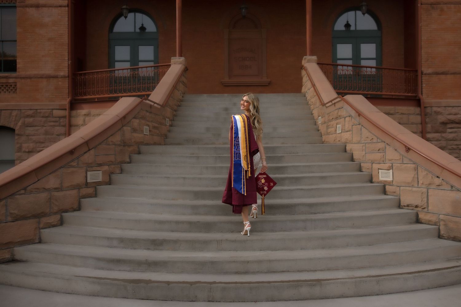 Graduate ascending historic Old Main building steps at Arizona State University wearing colorful graduation stoles and holding cap.