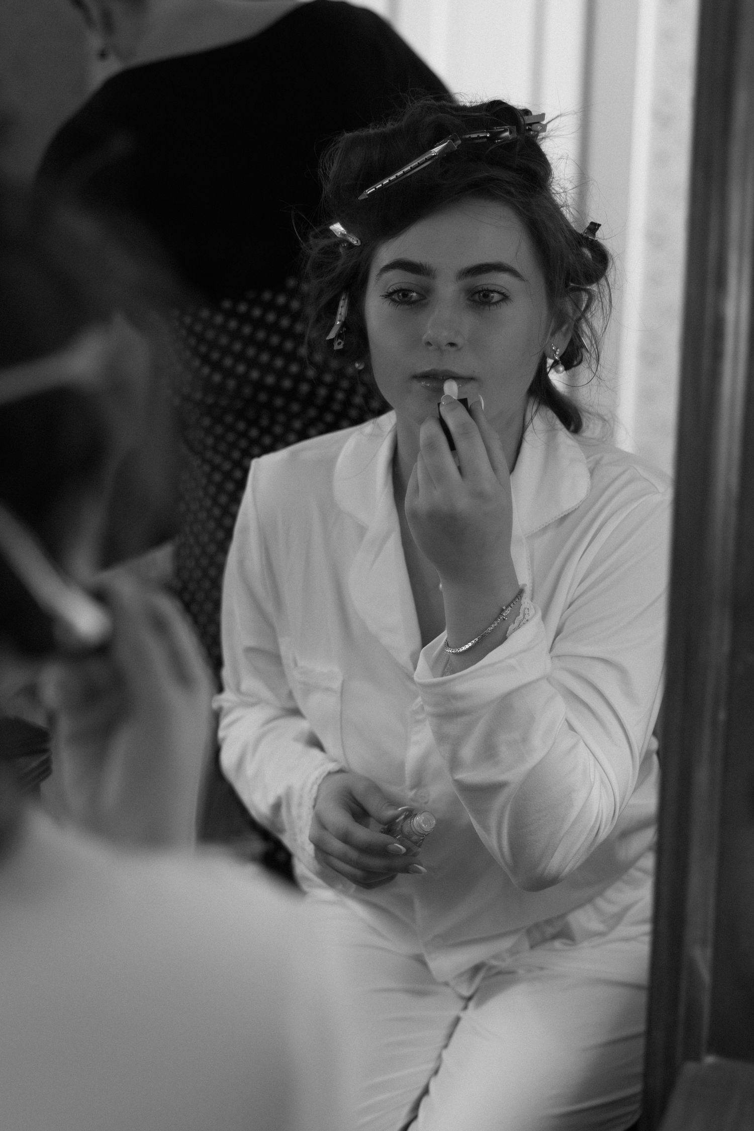Black and white photo of a bride applying makeup while looking in a mirror during wedding preparations.