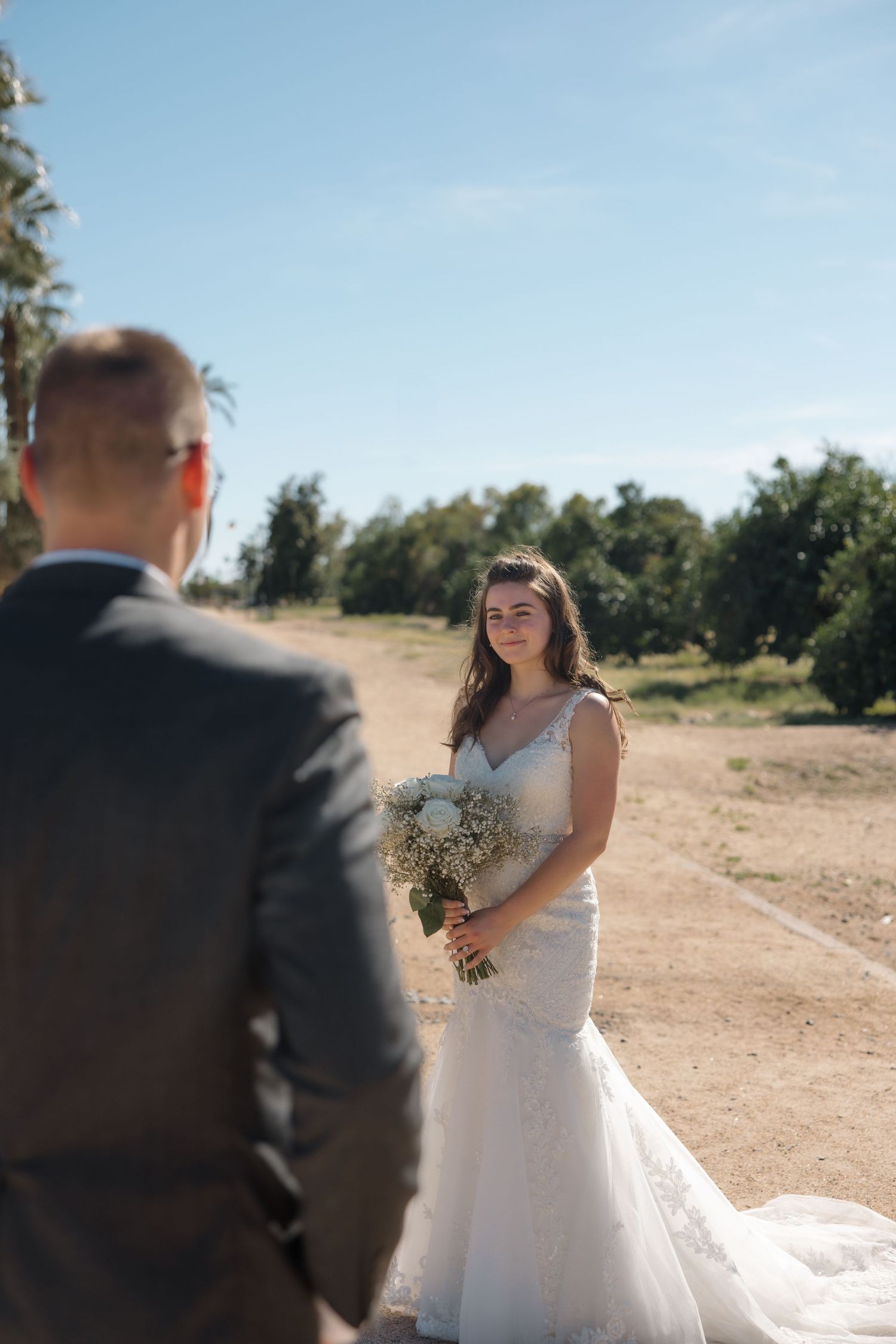 Bride in fitted lace wedding dress holding white bouquet faces groom on sandy outdoor path with palm trees.