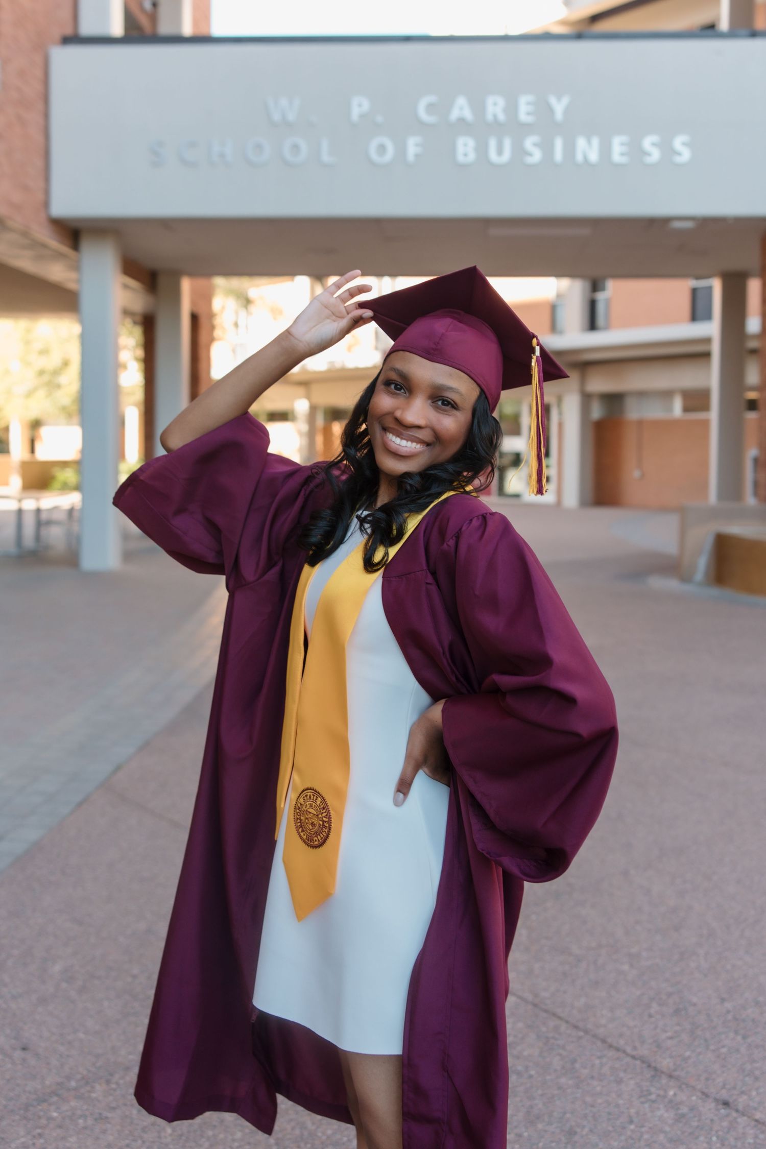 Smiling graduate in maroon cap and gown poses in front of W.P. Carey School of Business building.