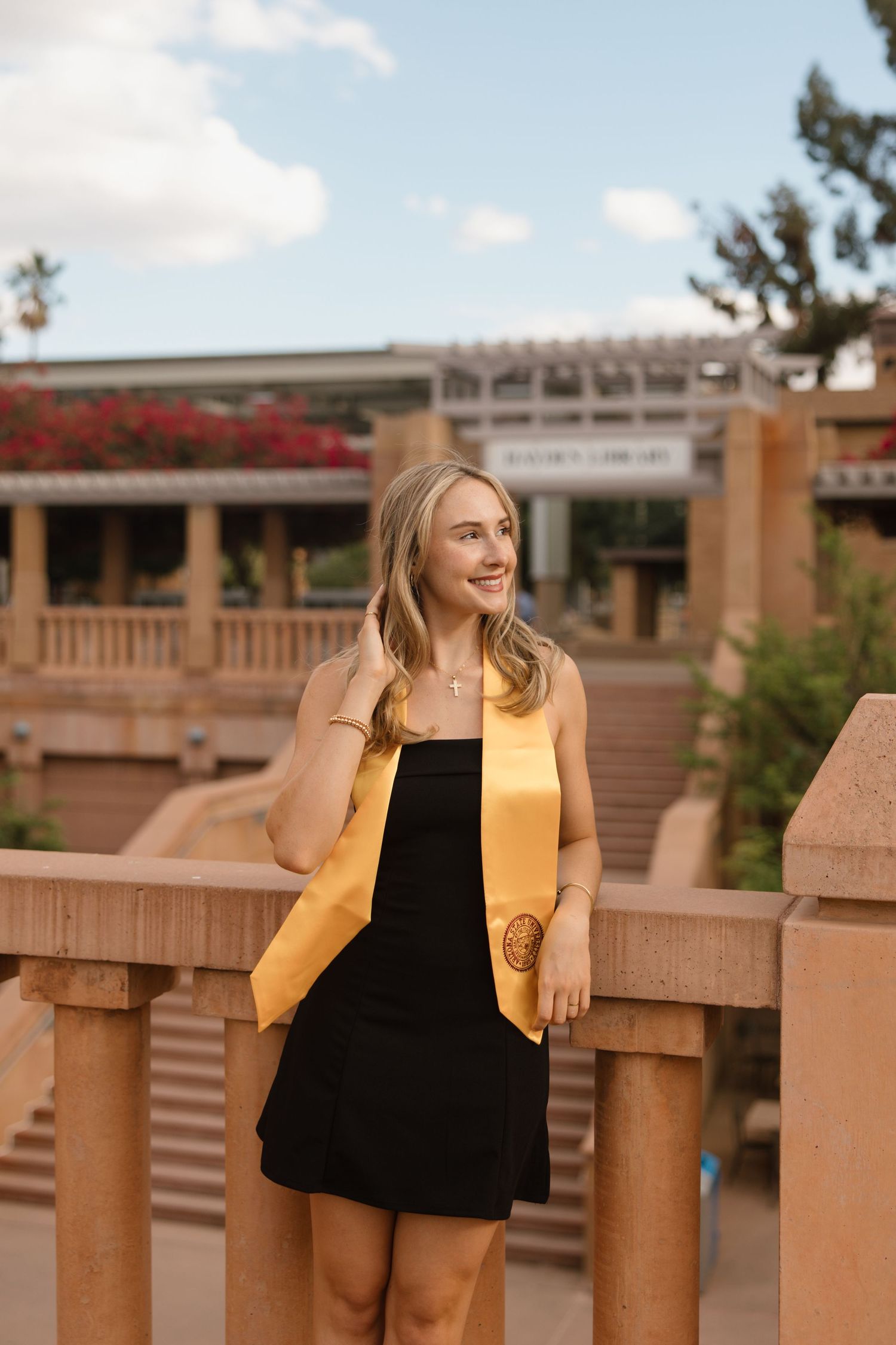 A smiling graduate in a black dress and gold sash poses outdoors on a campus bridge with red flowers in the background.
