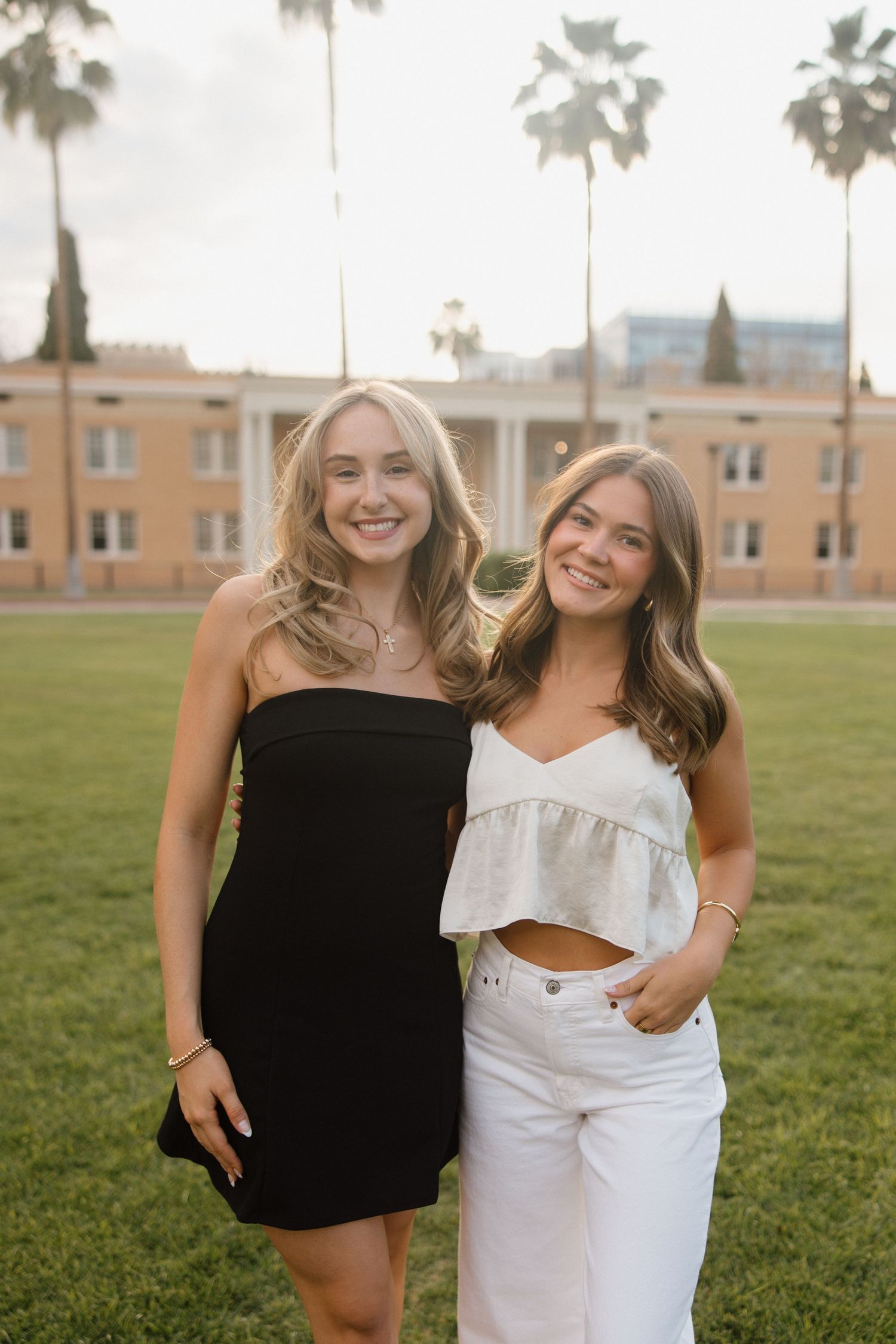 Two smiling young women posing outdoors on grass with palm trees and building behind them.