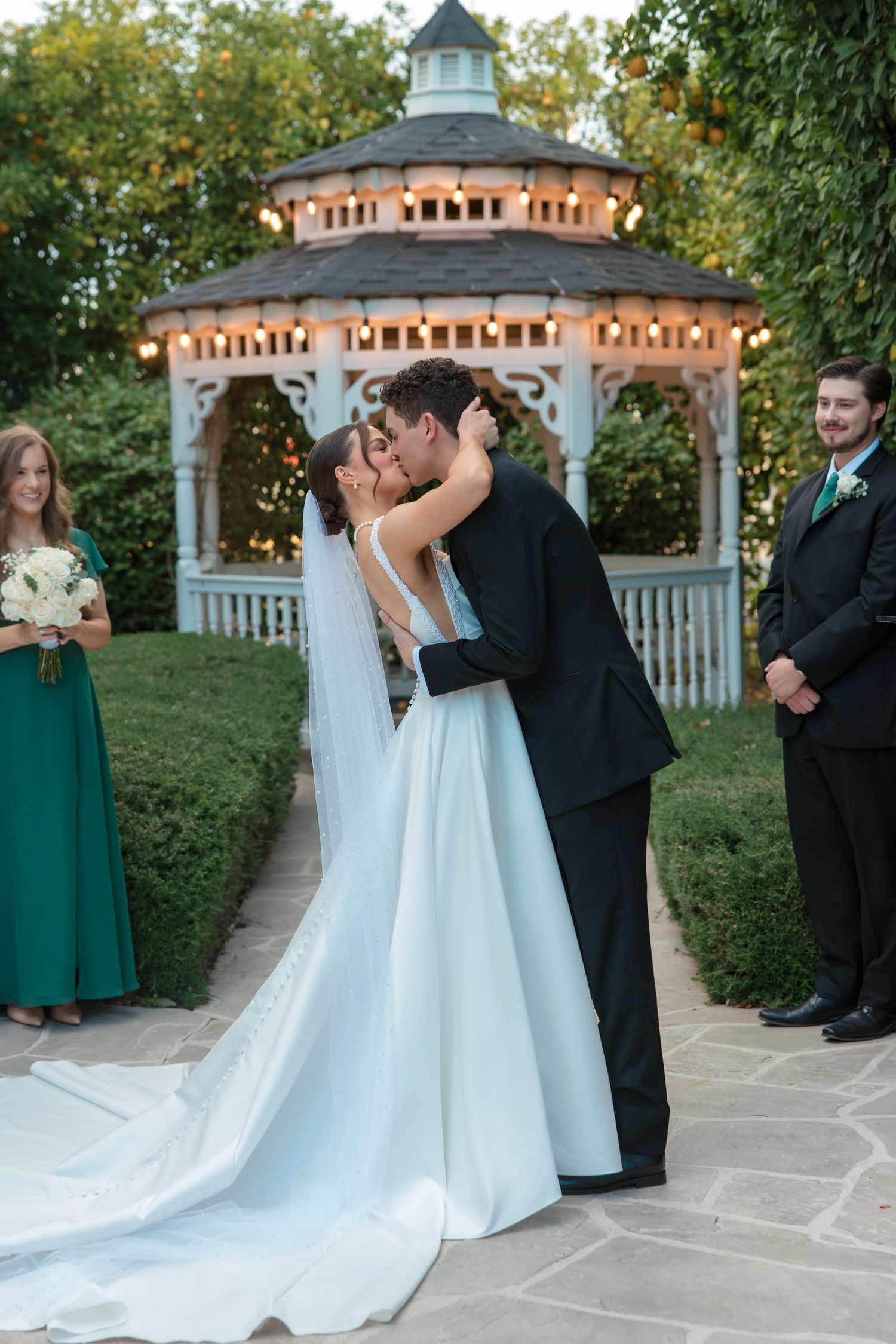Bride and groom share first kiss at outdoor gazebo wedding with bridesmaids in emerald green dresses.